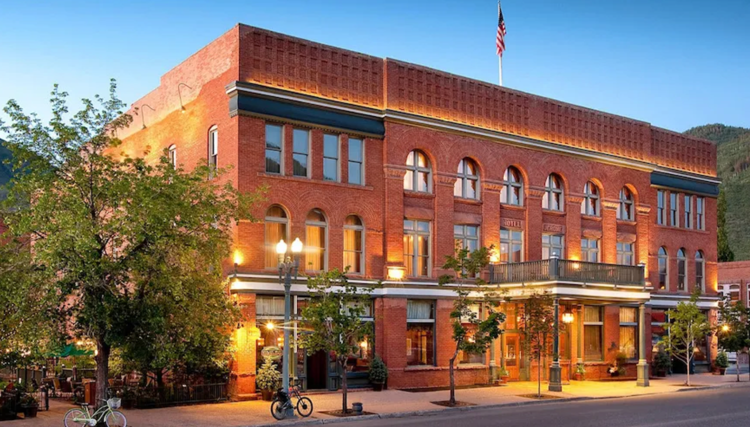 Historic red brick hotel building illuminated at dusk with American flag, arched windows, and tree-lined sidewalk.
