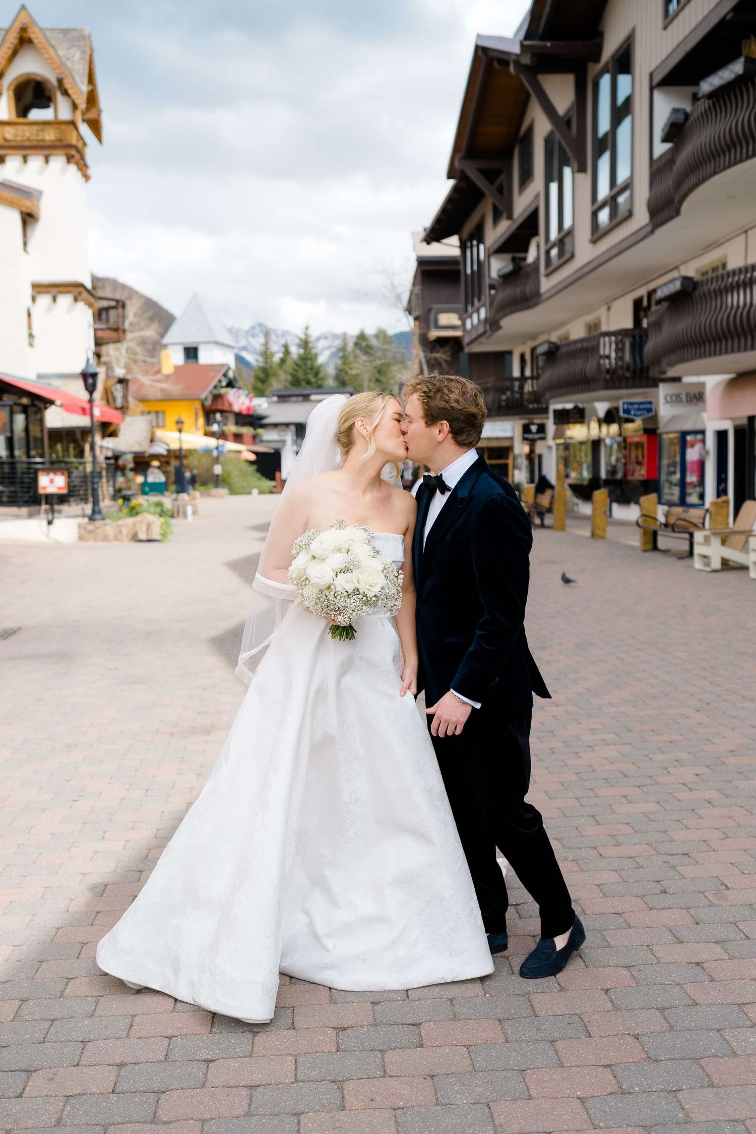 Bride and groom share a kiss while walking through a charming mountain village street, bride holding white bouquet.