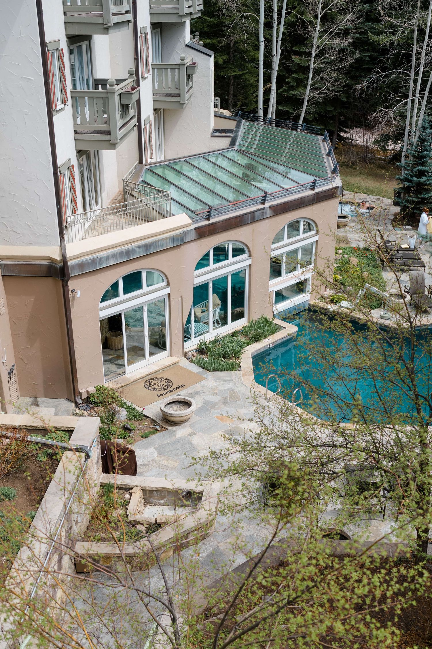 Aerial view of luxury home with glass conservatory, arched windows, stone patio, and turquoise pool surrounded by trees at Sonnenalp Hotel.