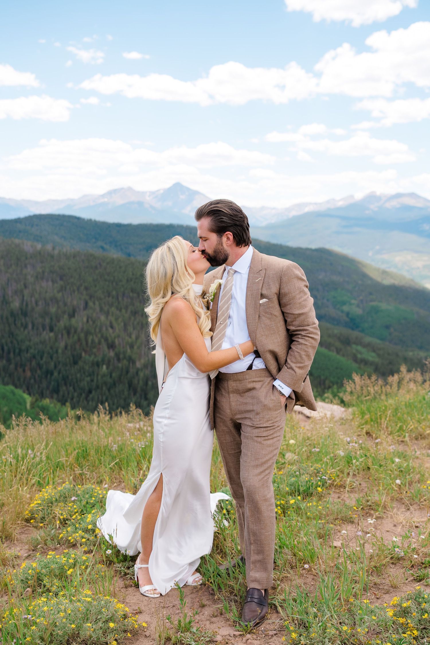 Bride and groom share a romantic kiss on a mountain overlook with lush green forests and blue sky backdrop.