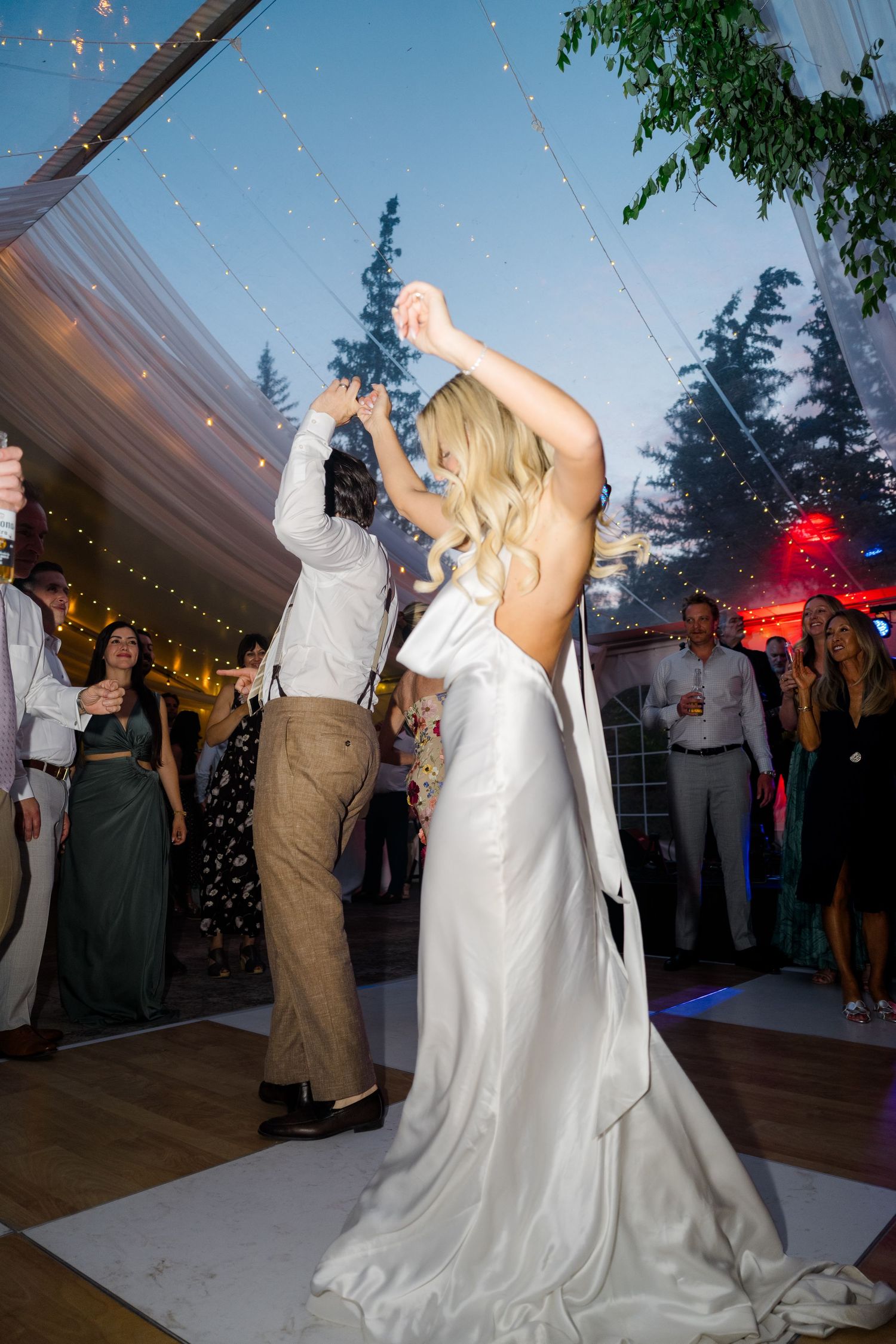 Bride in white backless gown dancing with groom at outdoor wedding reception under string lights at dusk.