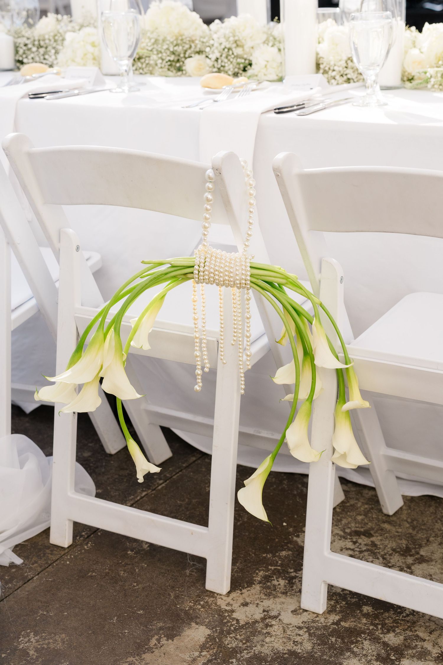 White folding chair decorated with calla lilies and pearl strands at an elegant wedding reception.