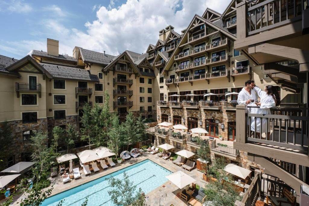 Four Seasons Vail Wedding Venue, Luxury mountain resort hotel with outdoor pool, sun loungers, and guests on balcony surrounded by trees.