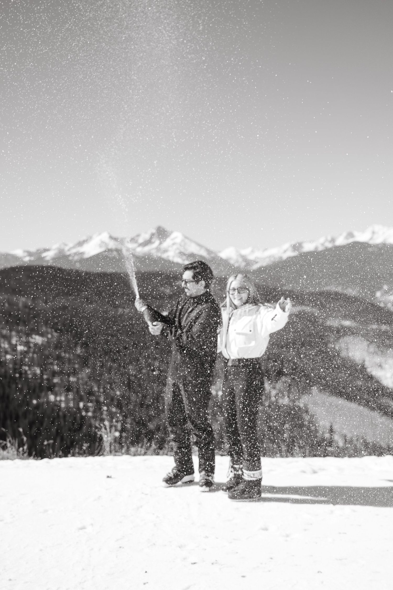 Couple celebrating engagement by popping champagne on snowy mountain with stunning winter landscape backdrop.