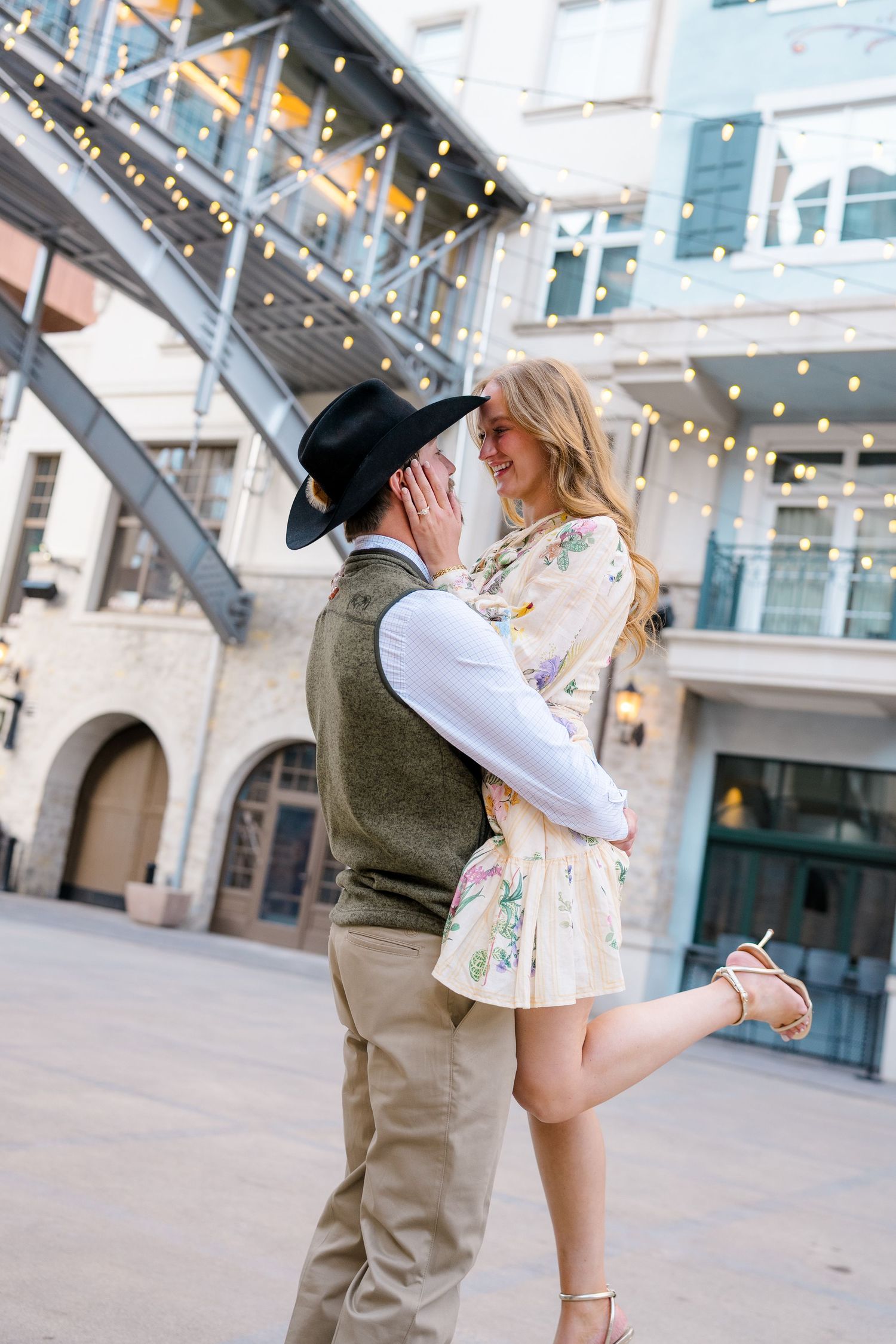 Couple sharing a romantic moment in an outdoor urban setting, woman in floral dress lifting leg as man in cowboy hat holds her close.