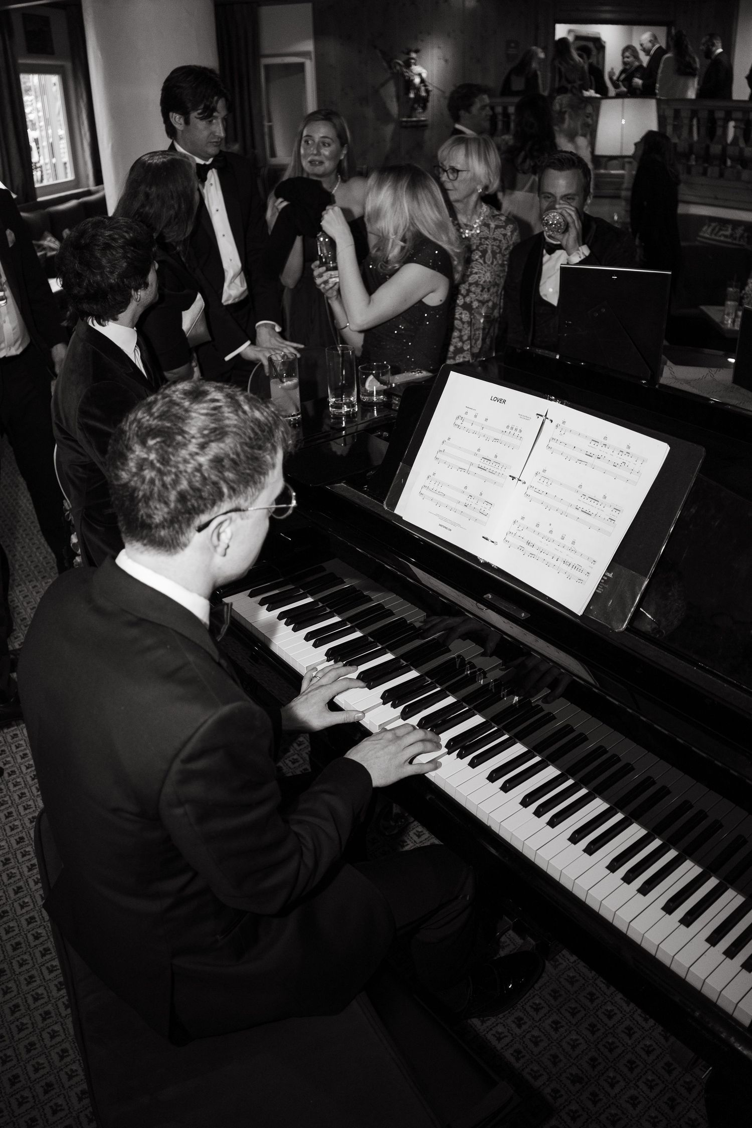 Black and white photo of a pianist playing grand piano at elegant party while guests socialize in background at Vail Sonnenalp Wedding.