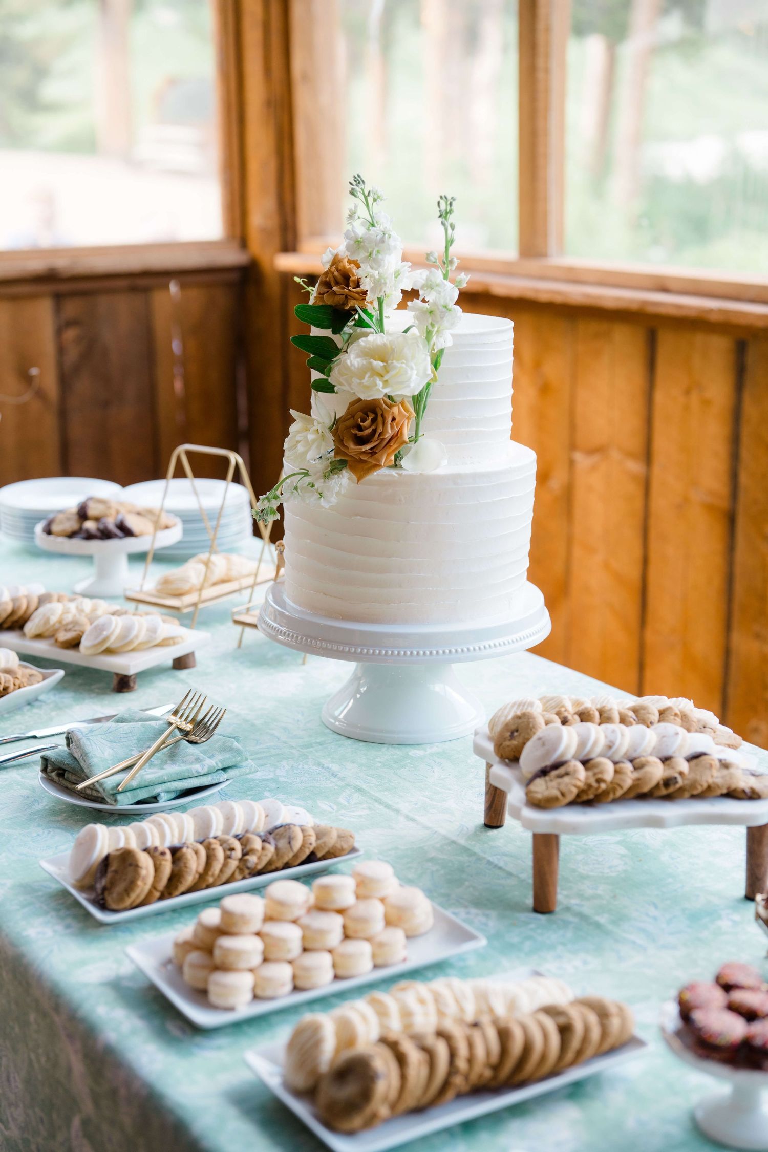 Elegant two-tier white wedding cake with floral topping surrounded by assorted cookies on a light blue tablecloth at Piney River Ranch.
