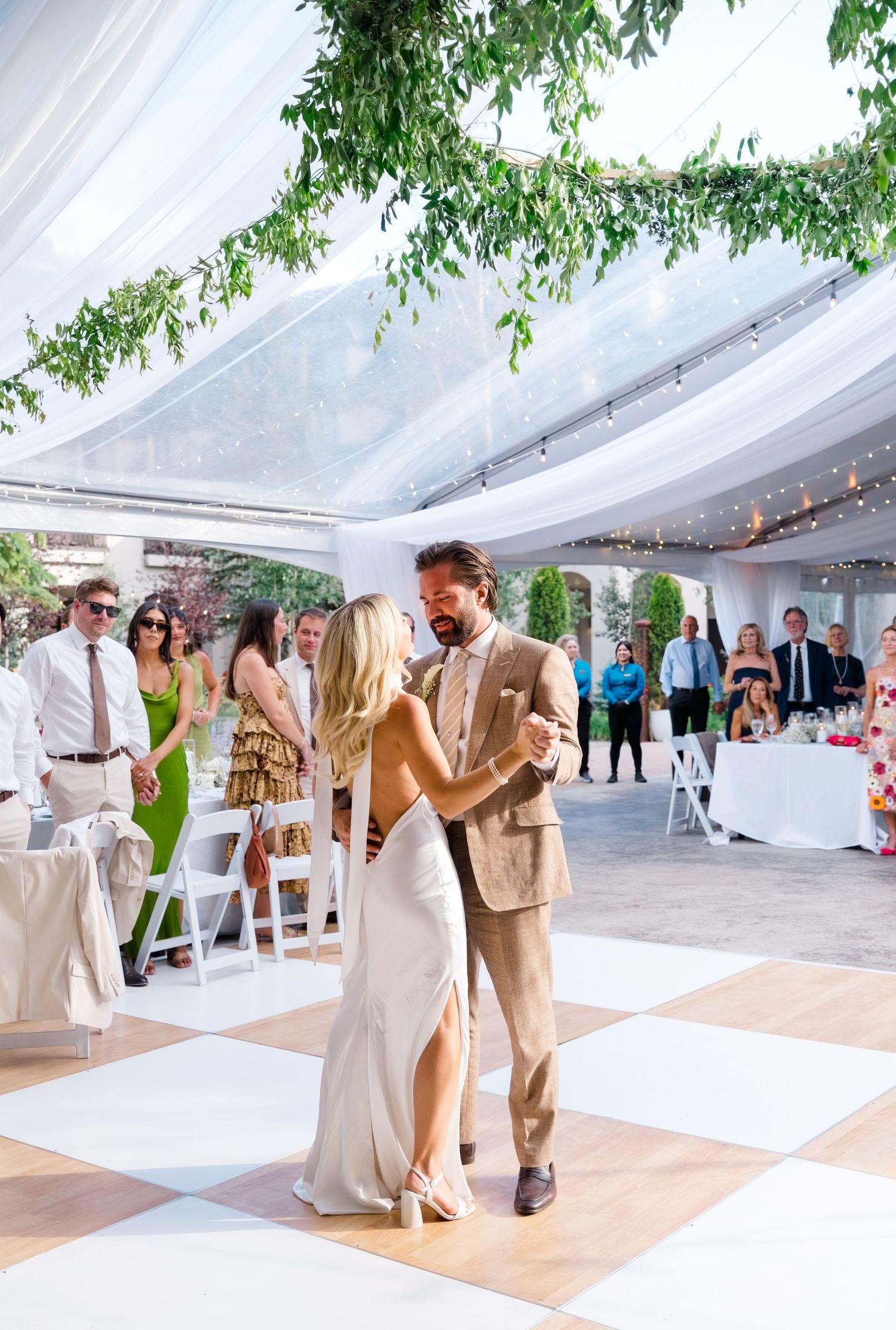 Couple shares first dance under a clear tent with greenery, guests watching on a checkered dance floor at an elegant wedding.