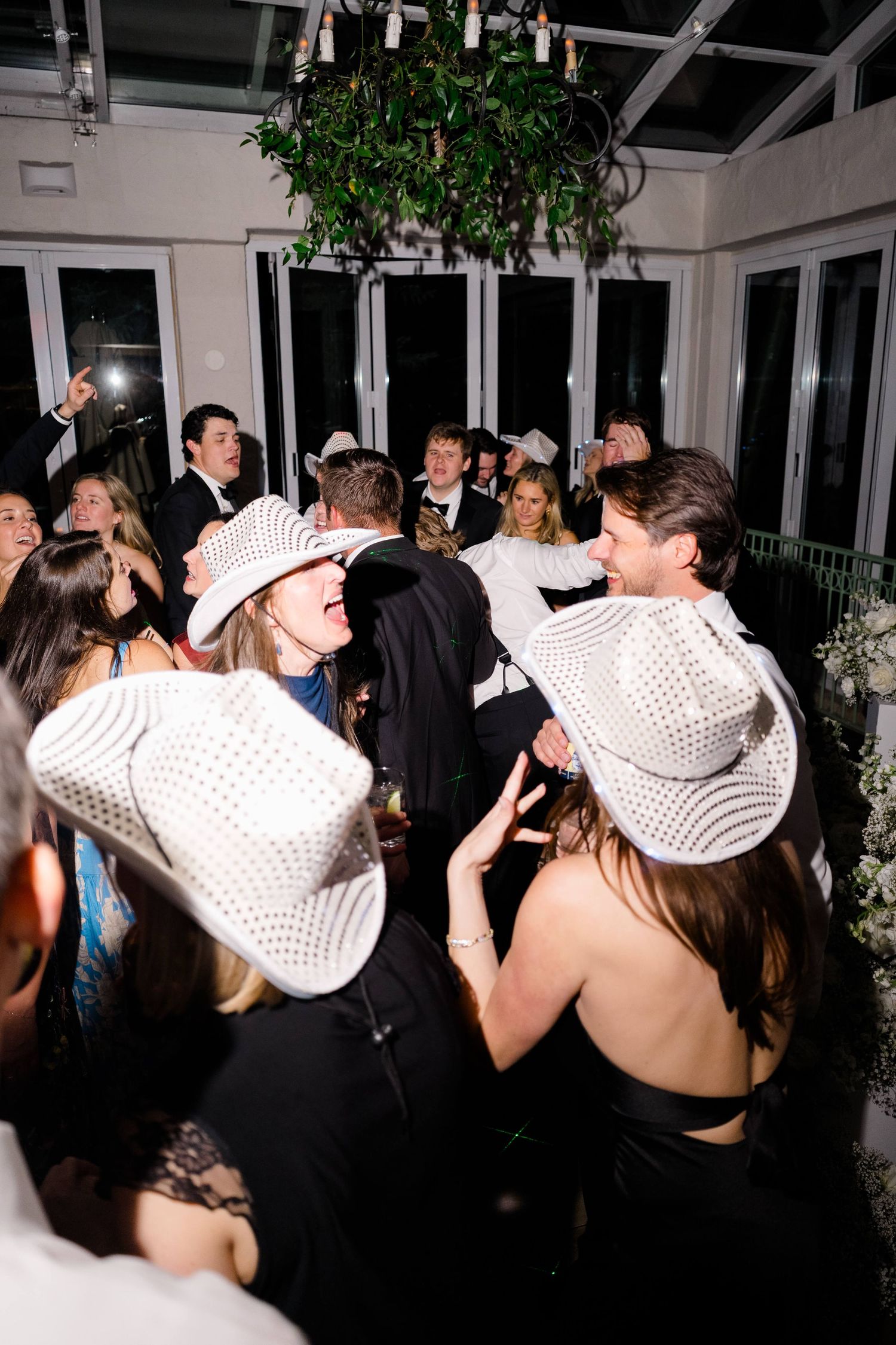 Guests dancing at a party wearing white cowboy hats in a decorated venue with chandelier.