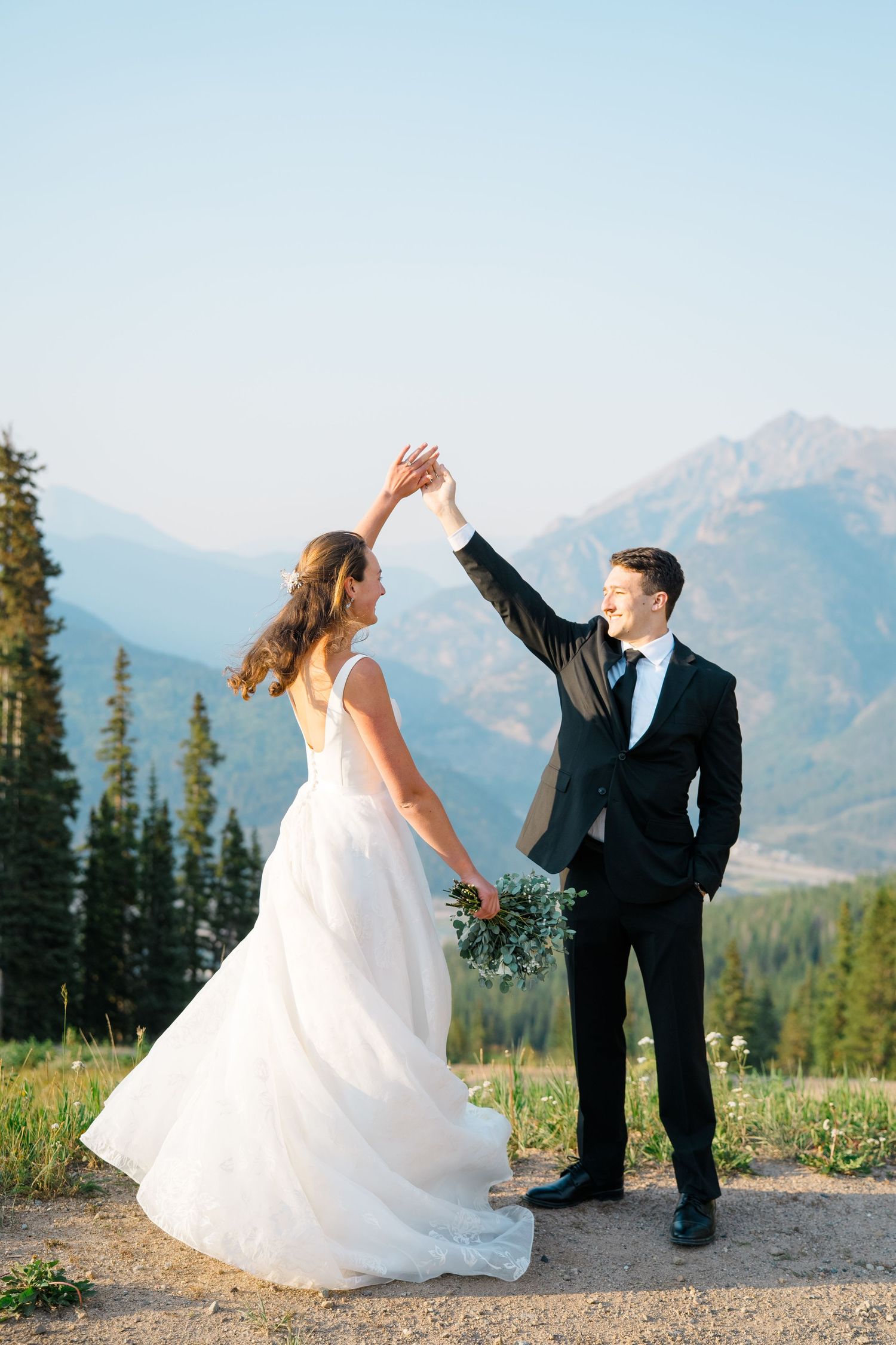 A couple dances playfully in a mountain meadow with scenic peaks in the background during their wedding photoshoot.