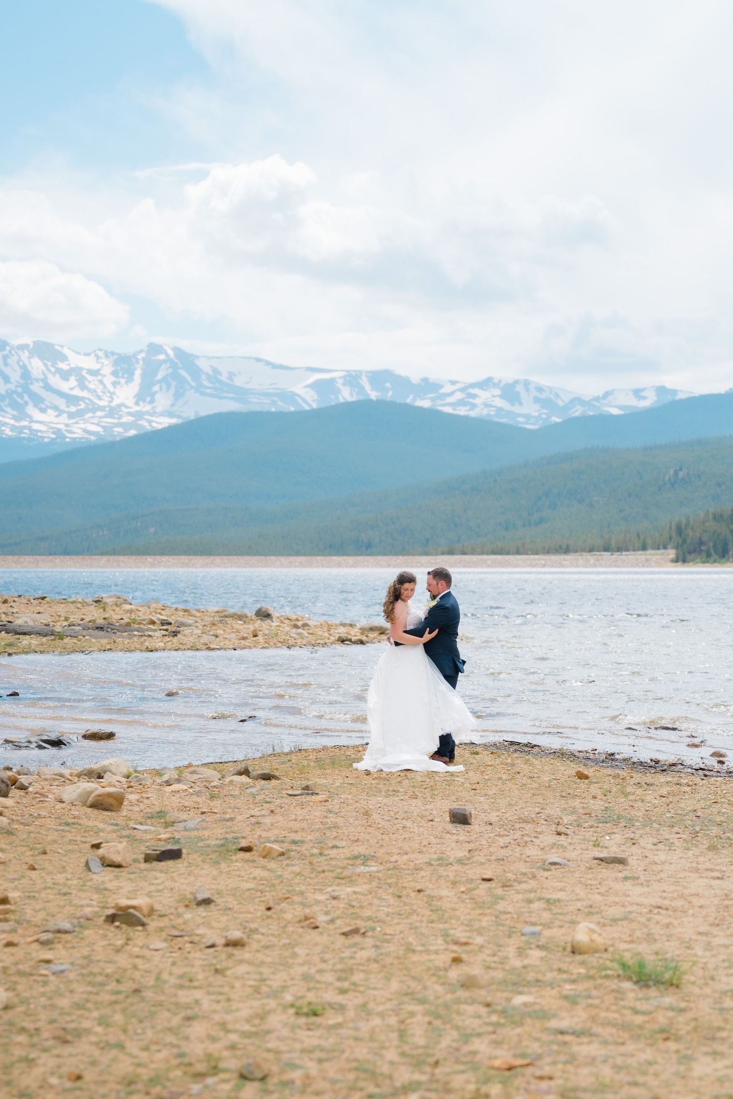 A wedding couple poses for photos on a beach with snow-capped mountains in the background.