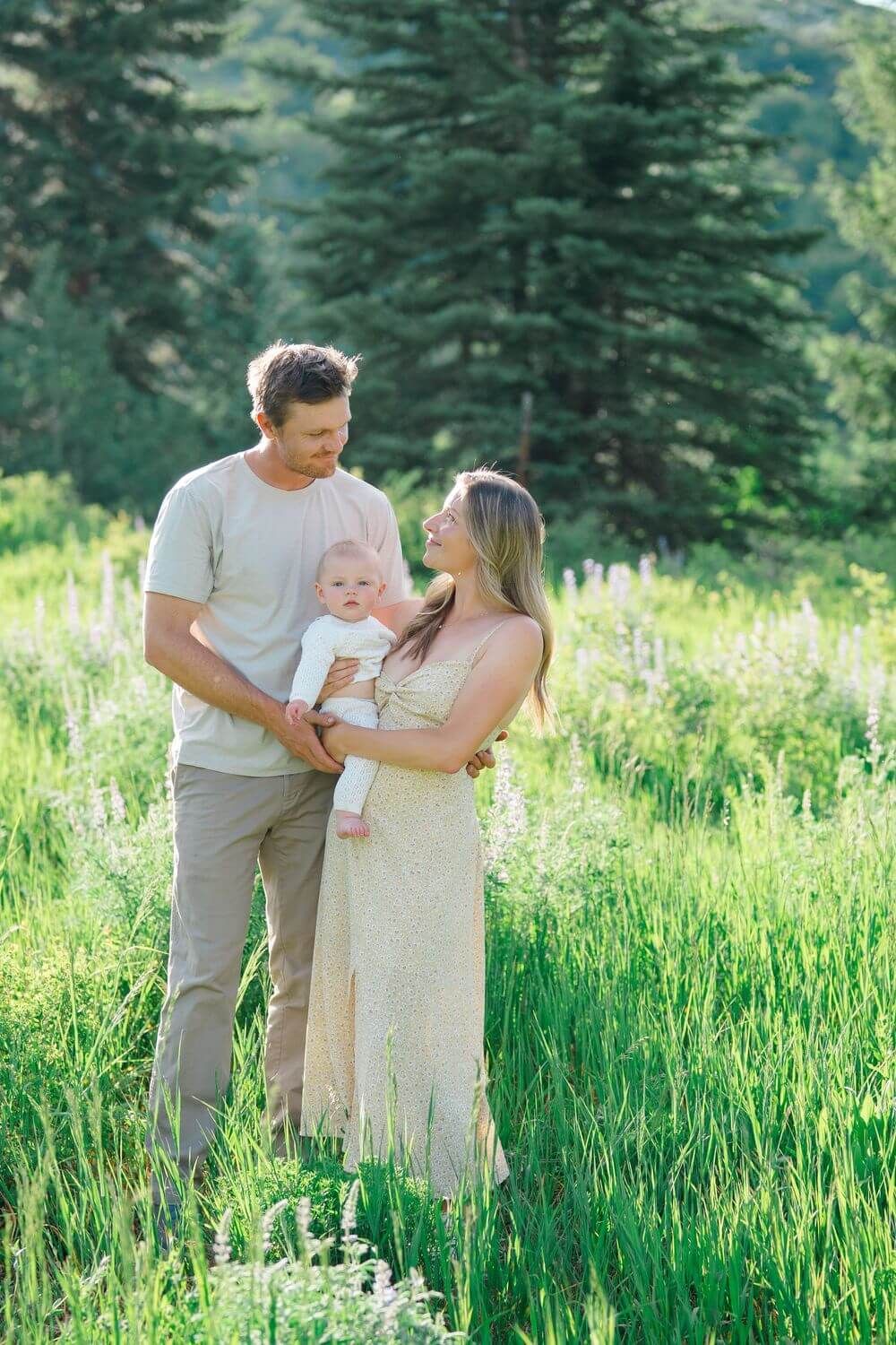 A couple with their baby stand together in a sunlit meadow with tall green grass and evergreen trees - vail family photo locations