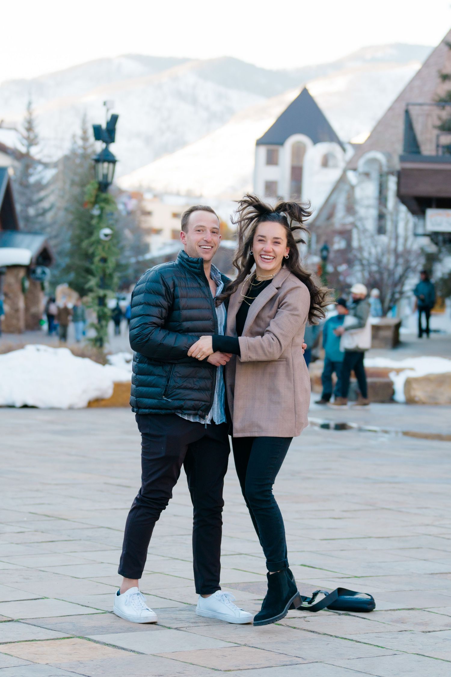 Two people share a joyful moment while posing together on a stone plaza in a winter mountain village setting.
