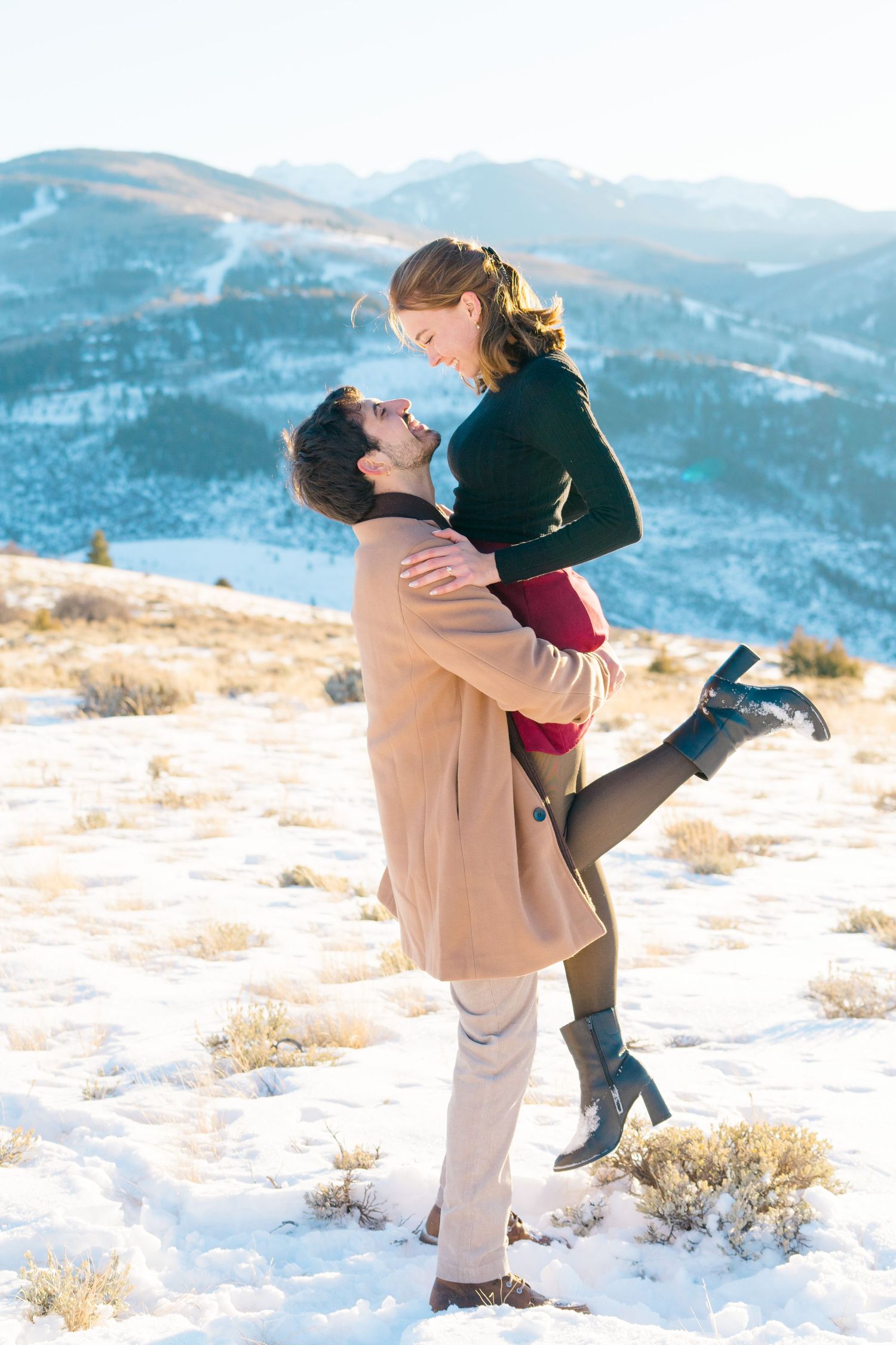 A romantic winter engagement photo shows a couple embracing on a snowy mountain overlook with scenic blue peaks in the distance.