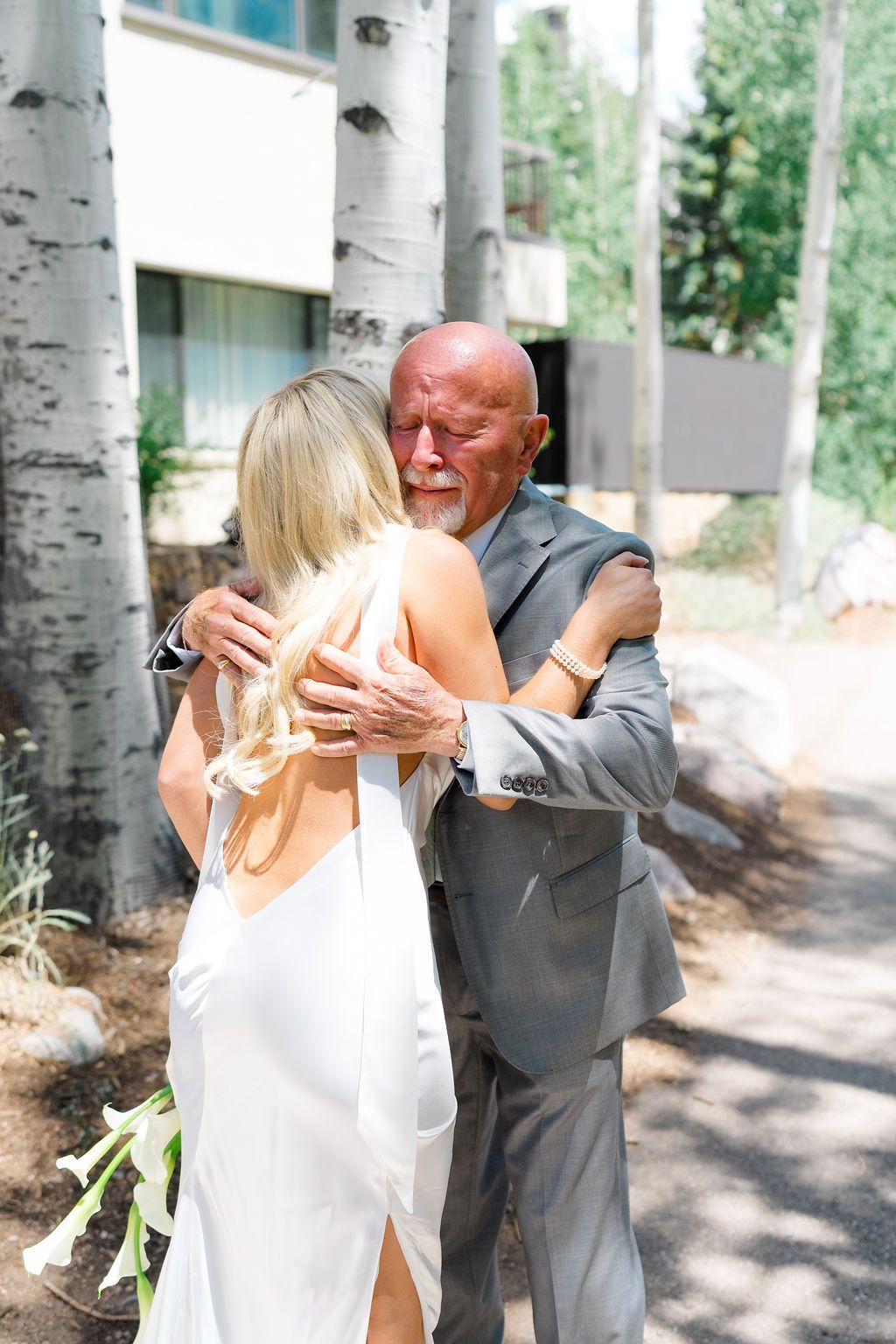Emotional father-daughter embrace at outdoor wedding, bride in white gown holding flowers near birch trees.