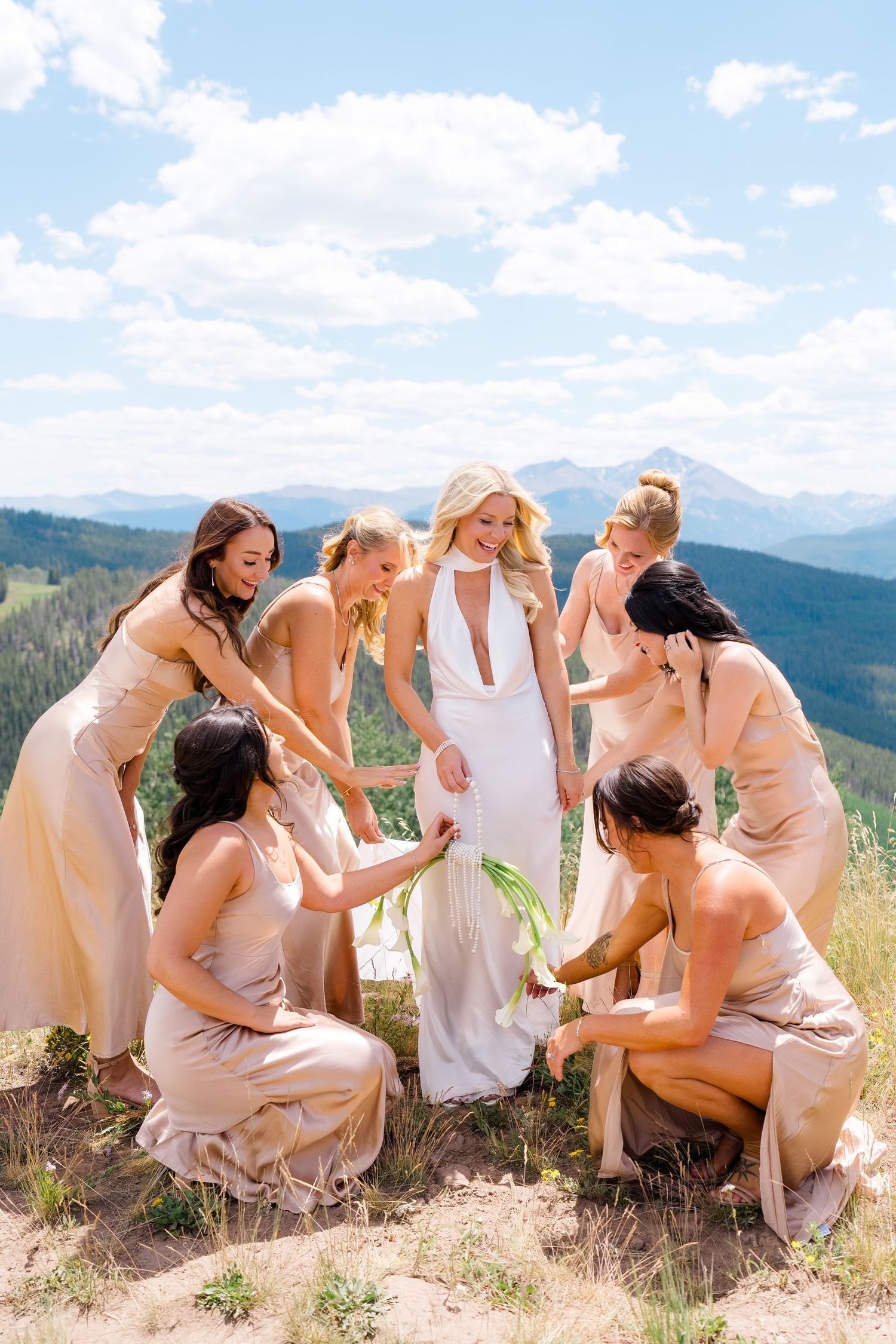 Bride in white gown surrounded by bridesmaids in champagne dresses on a mountain top with scenic views.