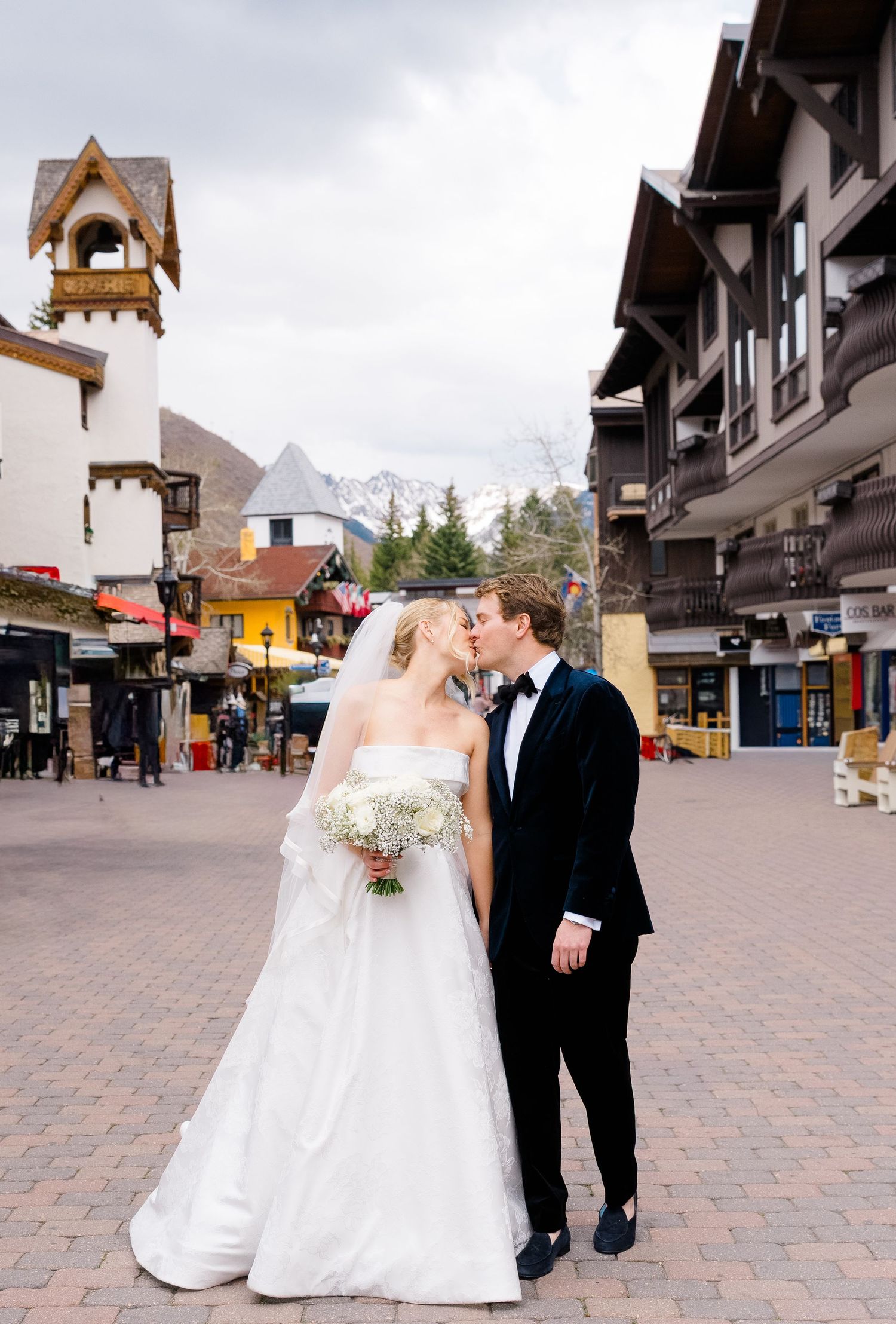 Bride and groom kissing in Vail Village plaza, she in white strapless gown with bouquet, he in black tuxedo, mountains behind at Sonnenalp.