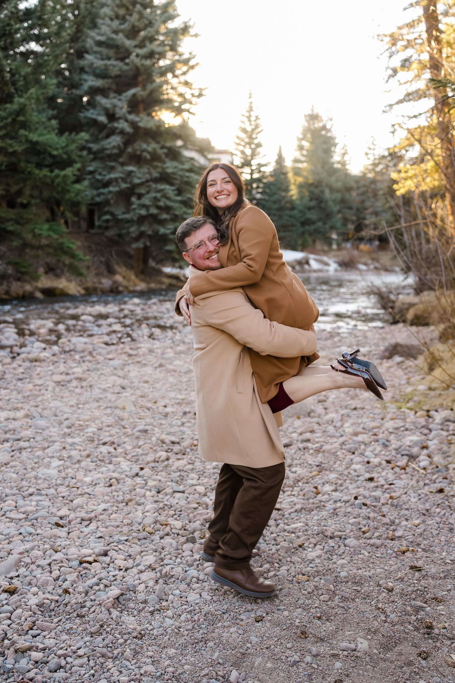 Man lifting smiling woman in a warm embrace on a rocky riverbank surrounded by pine trees at sunset.