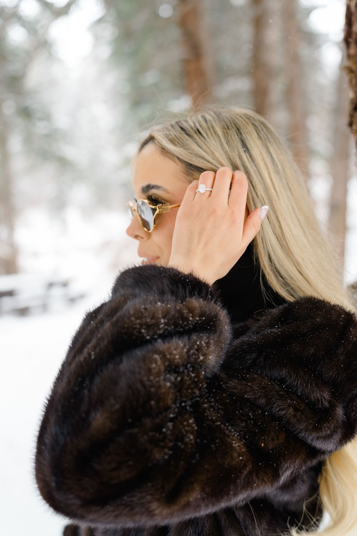 Blonde woman in dark mink fur coat and gold sunglasses posing in snowy winter forest setting.