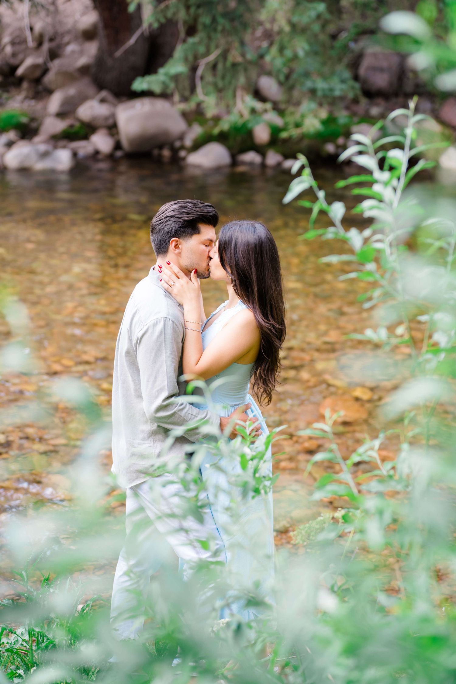 Following vail village proposal, a romantic couple shares a tender kiss by a tranquil stream surrounded by lush green foliage and leaves.