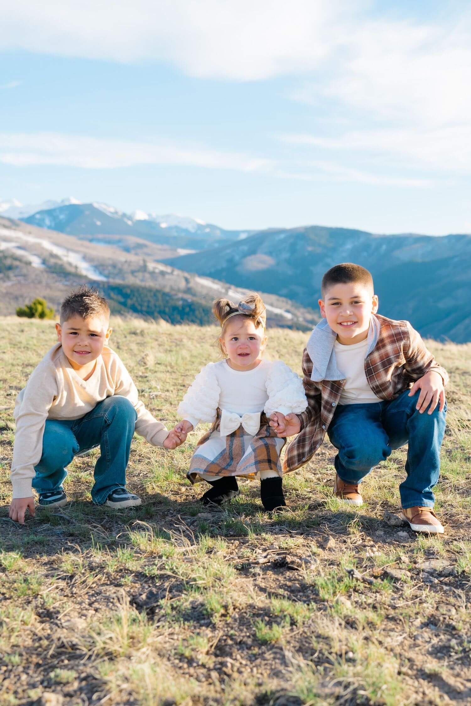 Four children sitting together on a grassy hillside with mountains visible in the background on a sunny day.