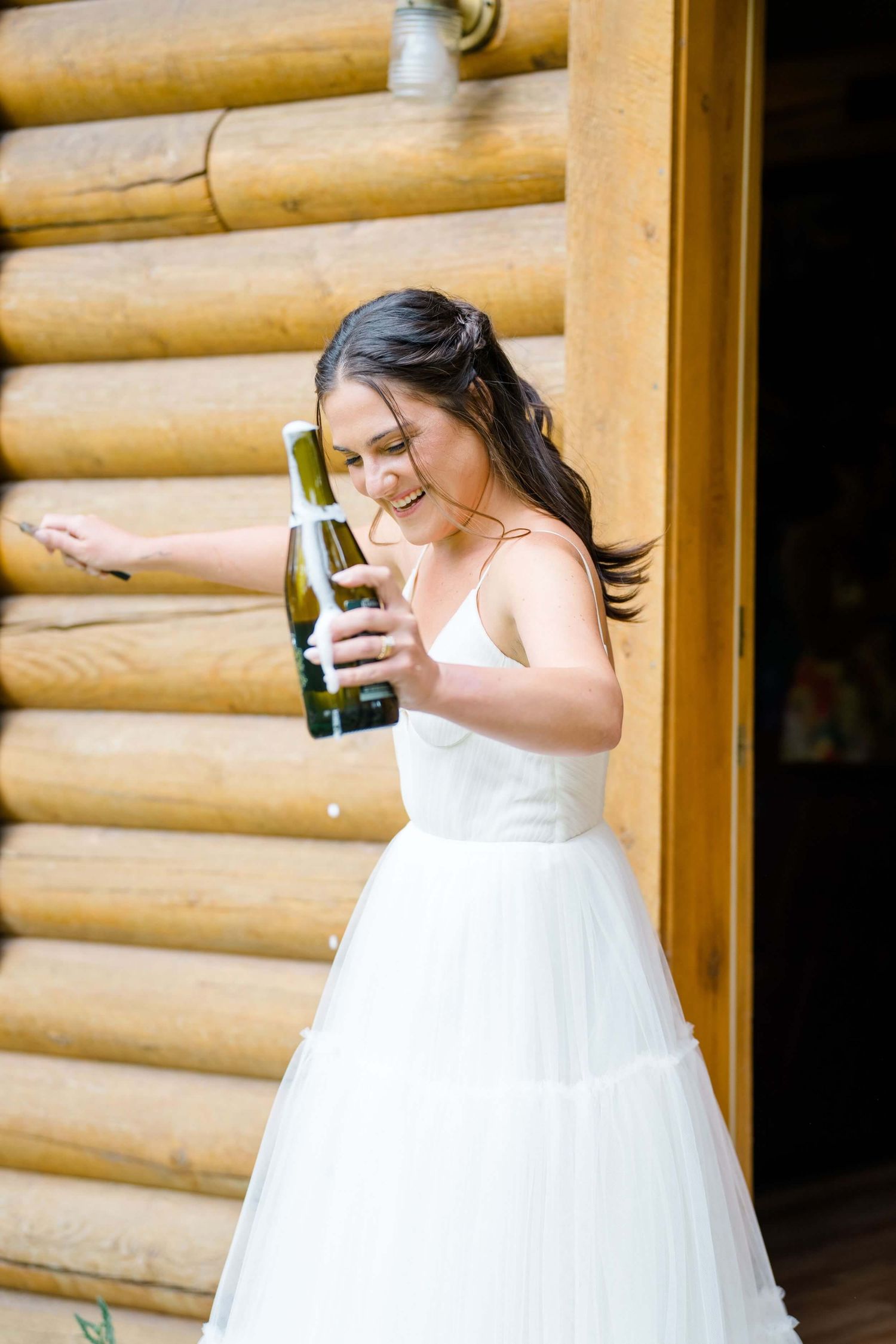 Camp Hale Wedding Bride Joyful moment captured at rustic log cabin wedding venue with bride holding champagne bottle.