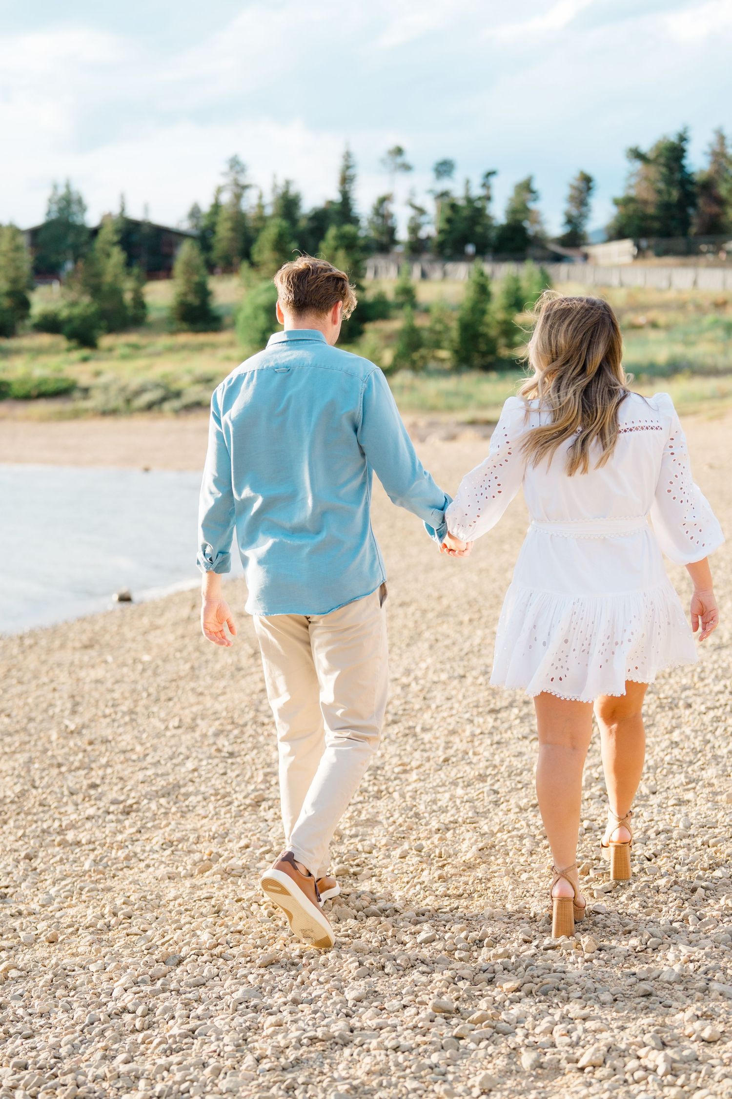 Two people in coordinating outfits walk hand in hand along a rocky beach with evergreen trees in the background at lake dillon.