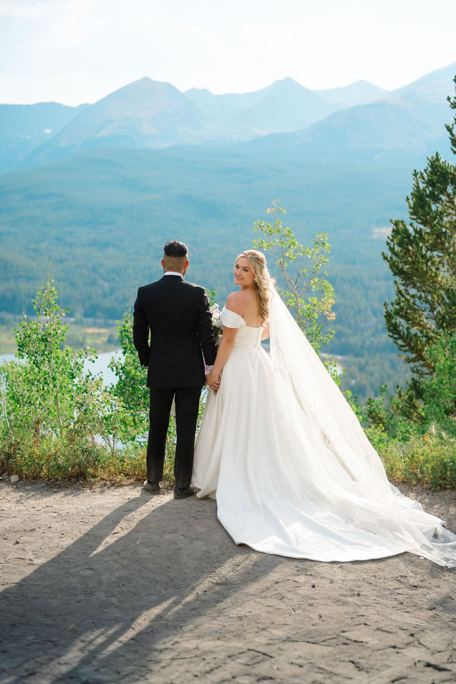A wedding couple stands at a scenic mountain overlook with a flowing white dress and dramatic shadows.