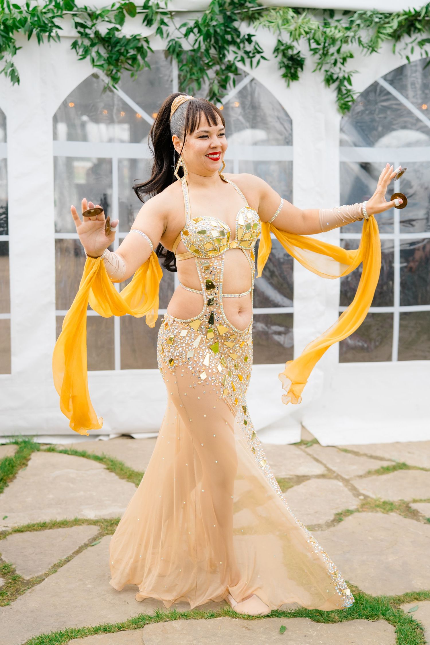 Dancer performing in sparkly gold dress with flowing yellow scarves against white greenhouse backdrop.