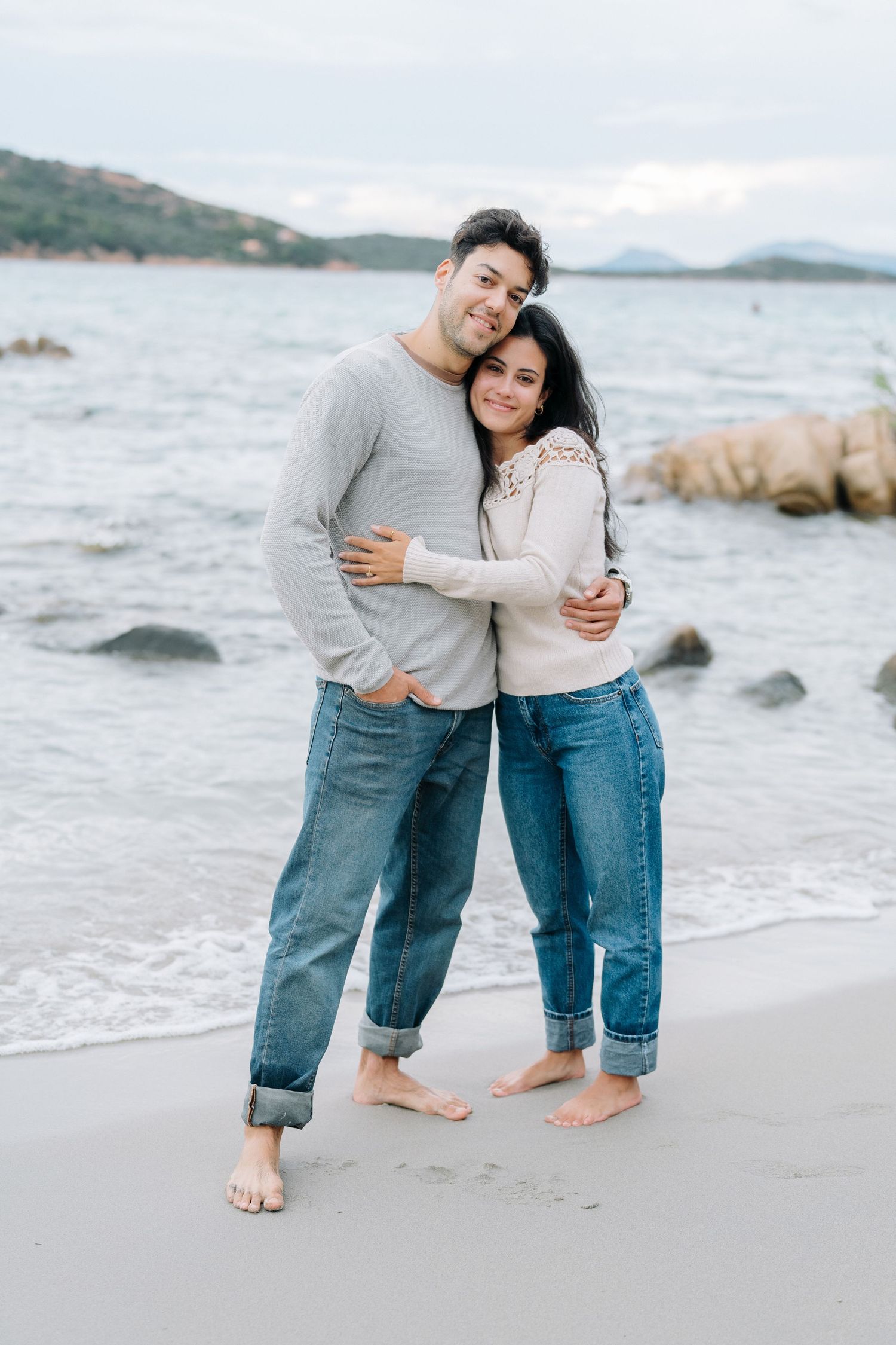 A couple embraces while standing barefoot on a rocky beach with mountains and water in the background.