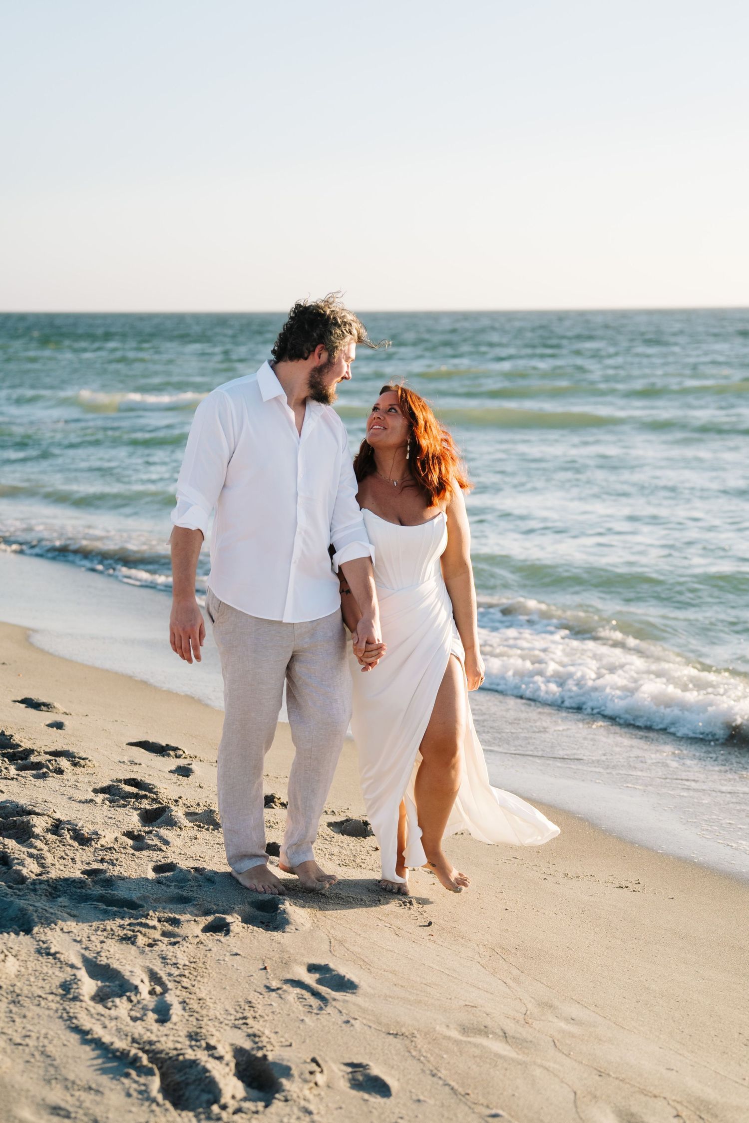 A couple in white attire walking hand in hand along a beach at sunset with waves crashing in the background.