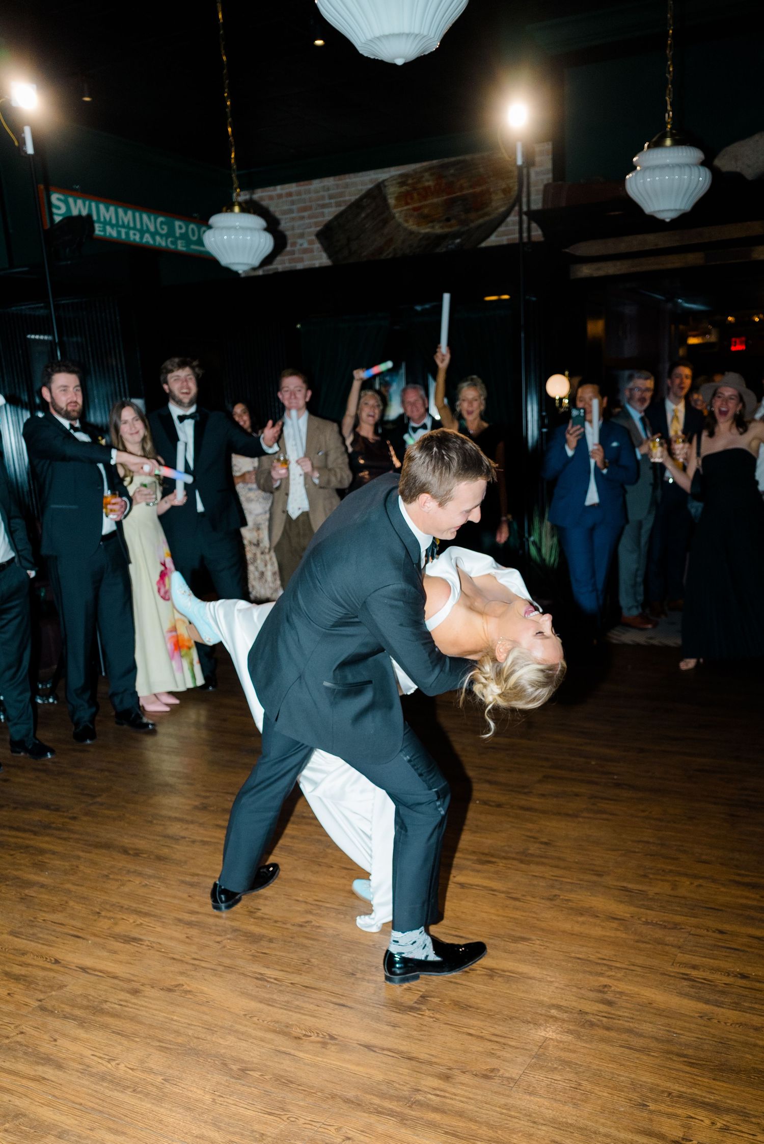 A wedding couple performs a dramatic dance dip on a wooden dance floor while guests cheer in the background.