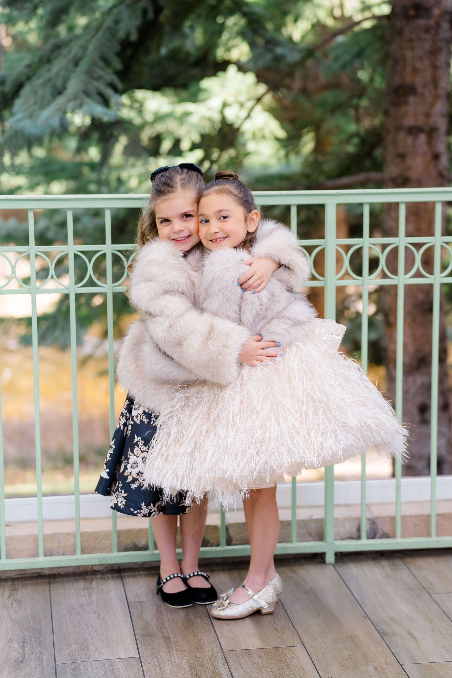 Two young girls in fluffy white fur coats hugging and smiling on a wooden deck with green railing.