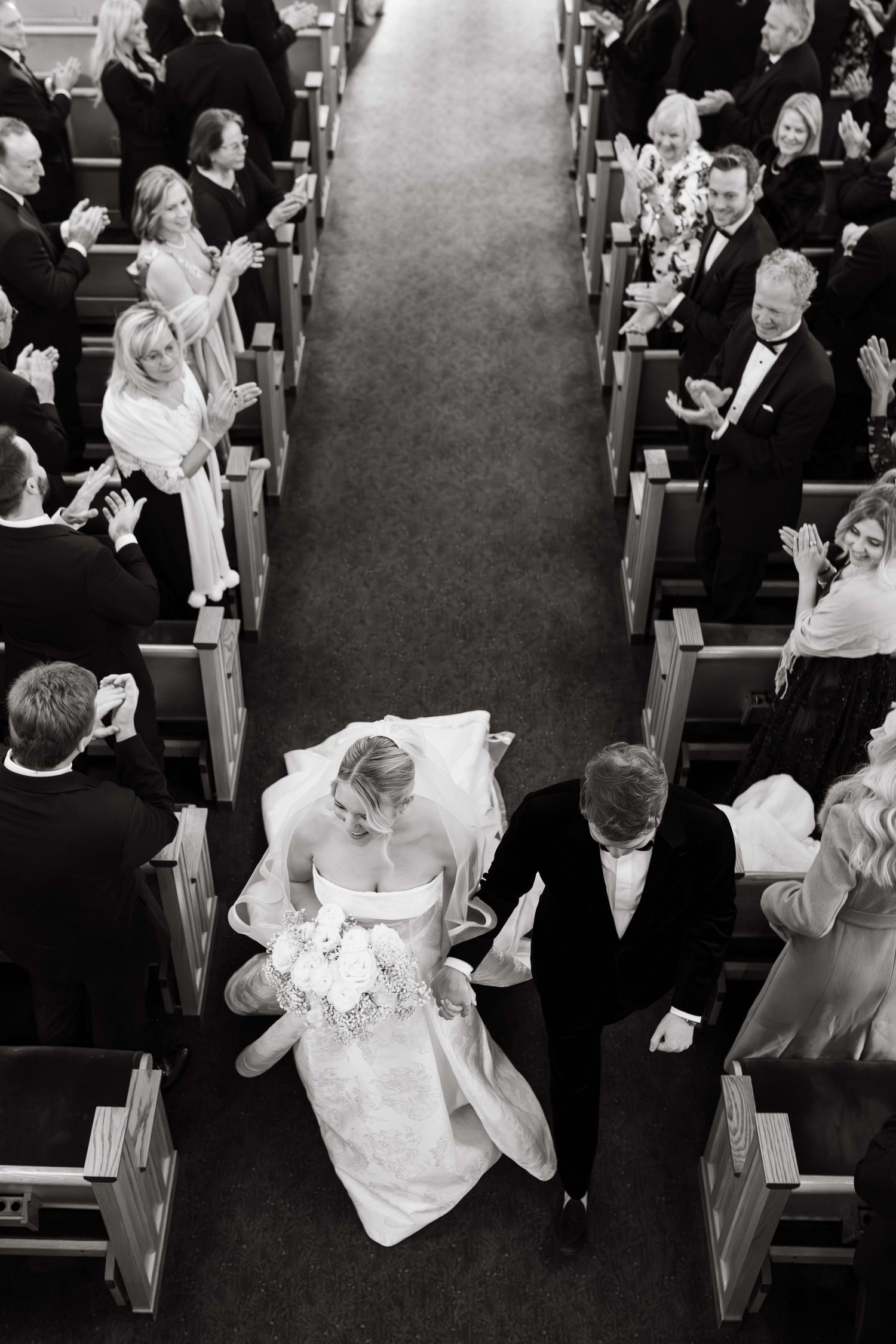 Aerial black and white photo of bride and groom walking down church aisle with guests applauding.