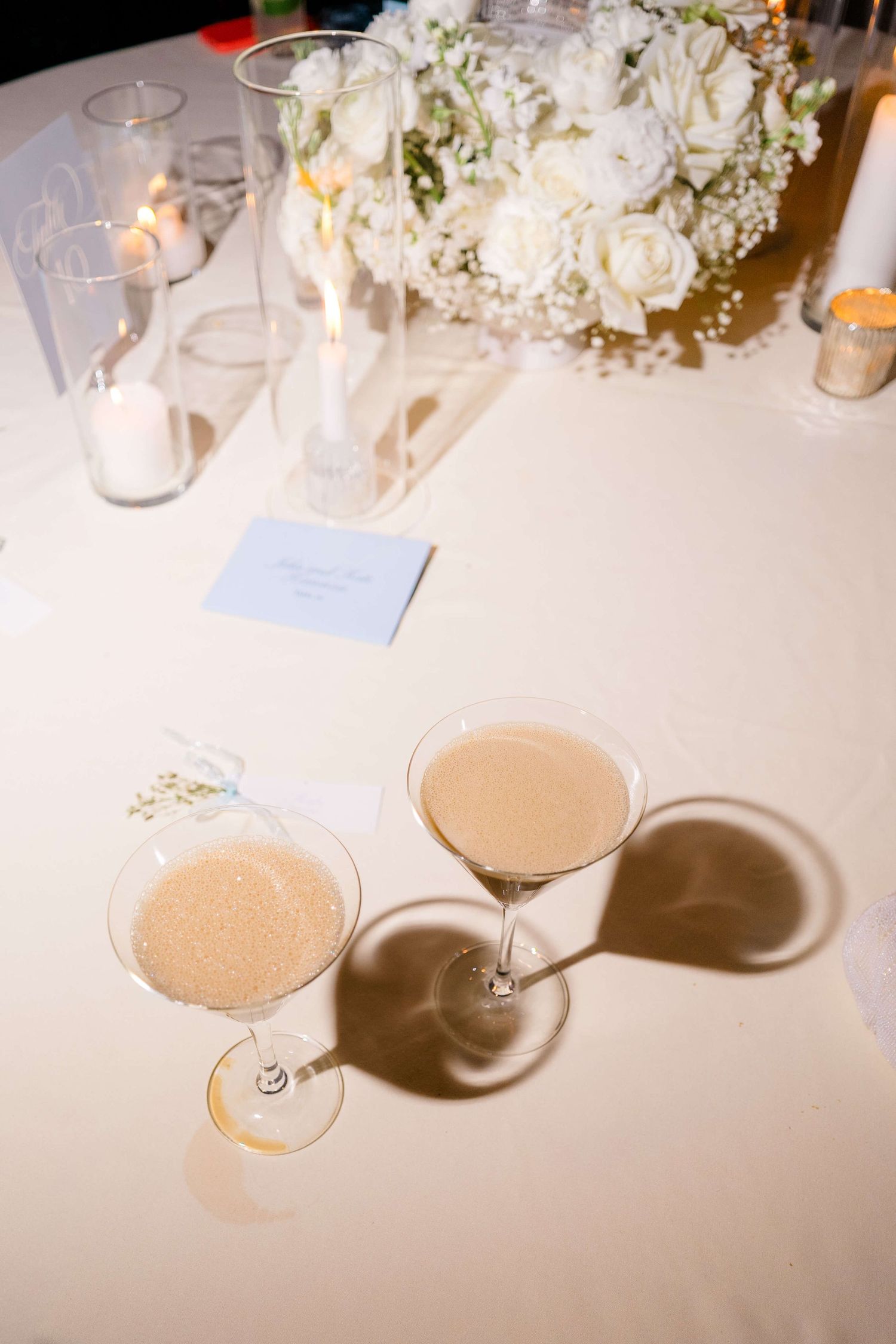 Two champagne cocktails on a white wedding reception table with candles and white roses centerpiece.