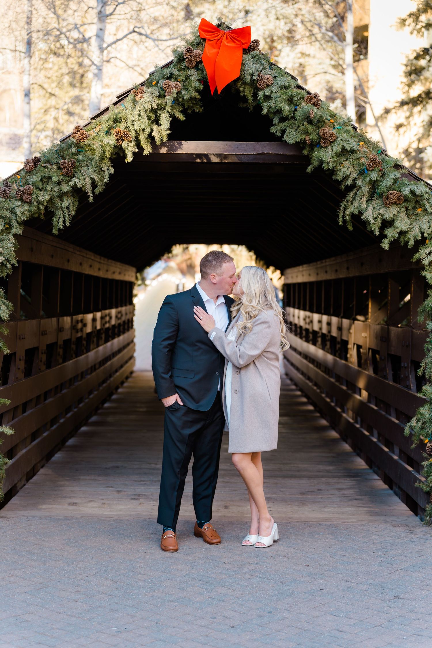 A romantic couple shares an intimate moment on a wooden covered bridge surrounded by evergreen trees.