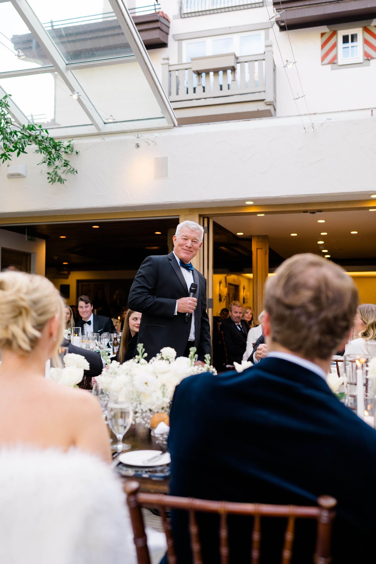 Man in tuxedo giving a wedding toast speech to bride and groom at elegant indoor reception venue.