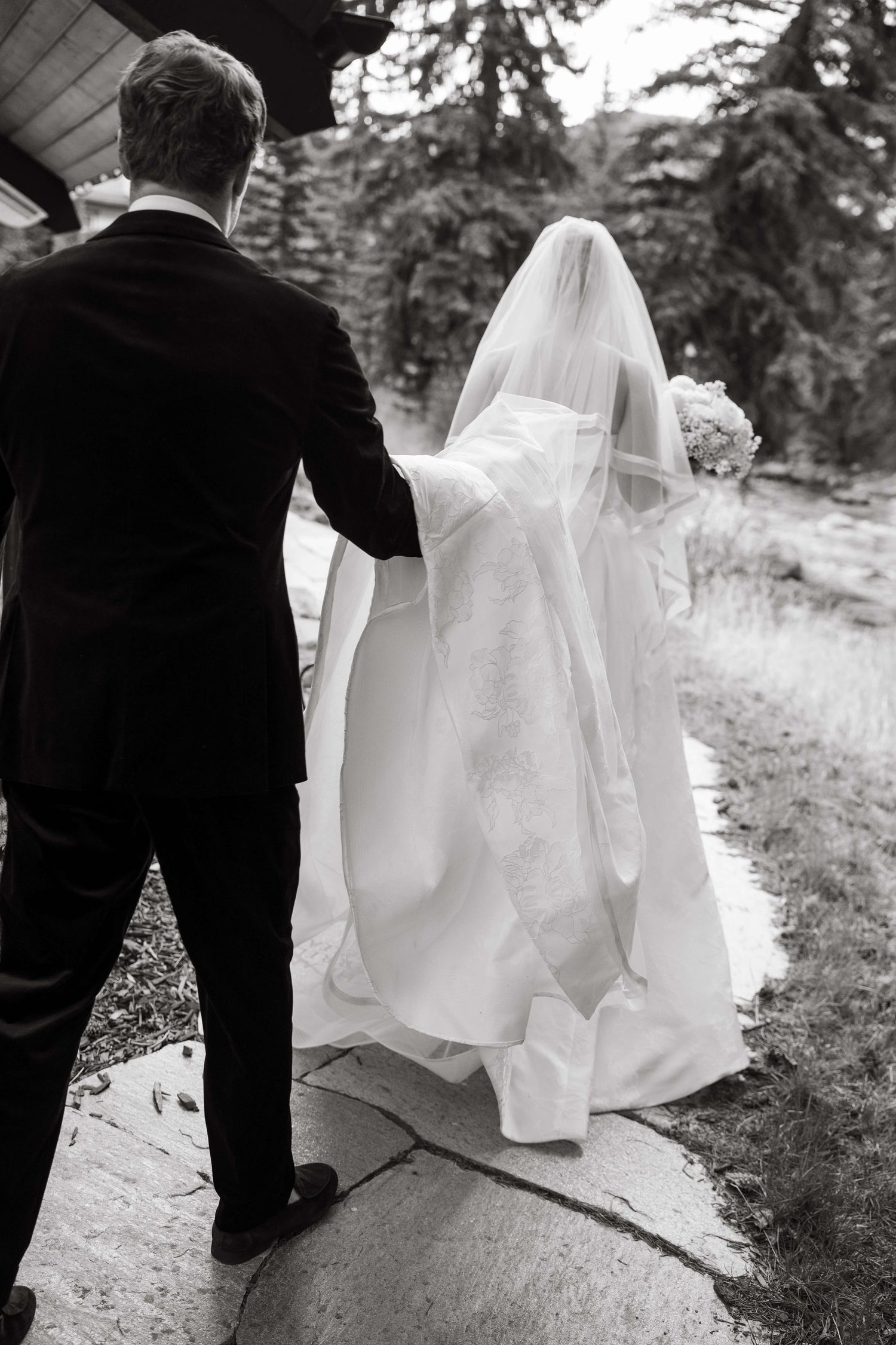 Black and white photo of bride and groom walking away, bride in flowing gown with veil and bouquet.