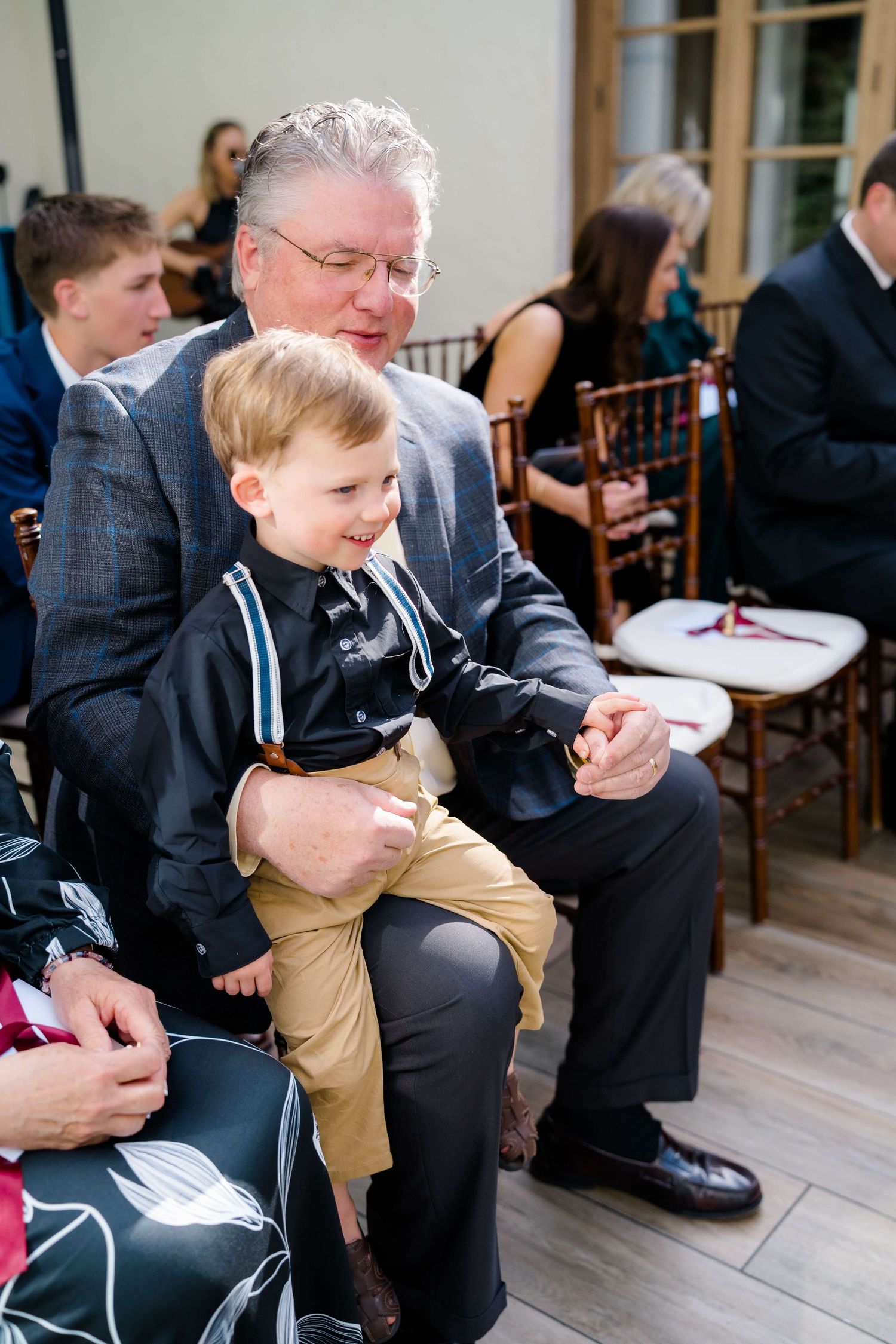 Grandfather holding young boy in suspenders at a wedding ceremony, both smiling and dressed in formal attire.