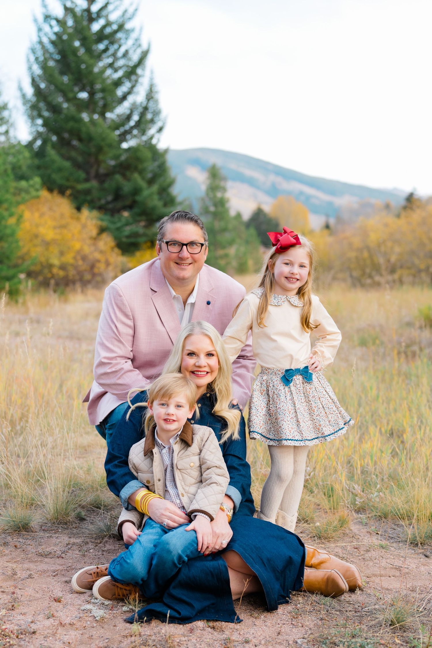 A family in coordinating pink and blue outfits poses together outdoors in a natural field setting with mountains in background.