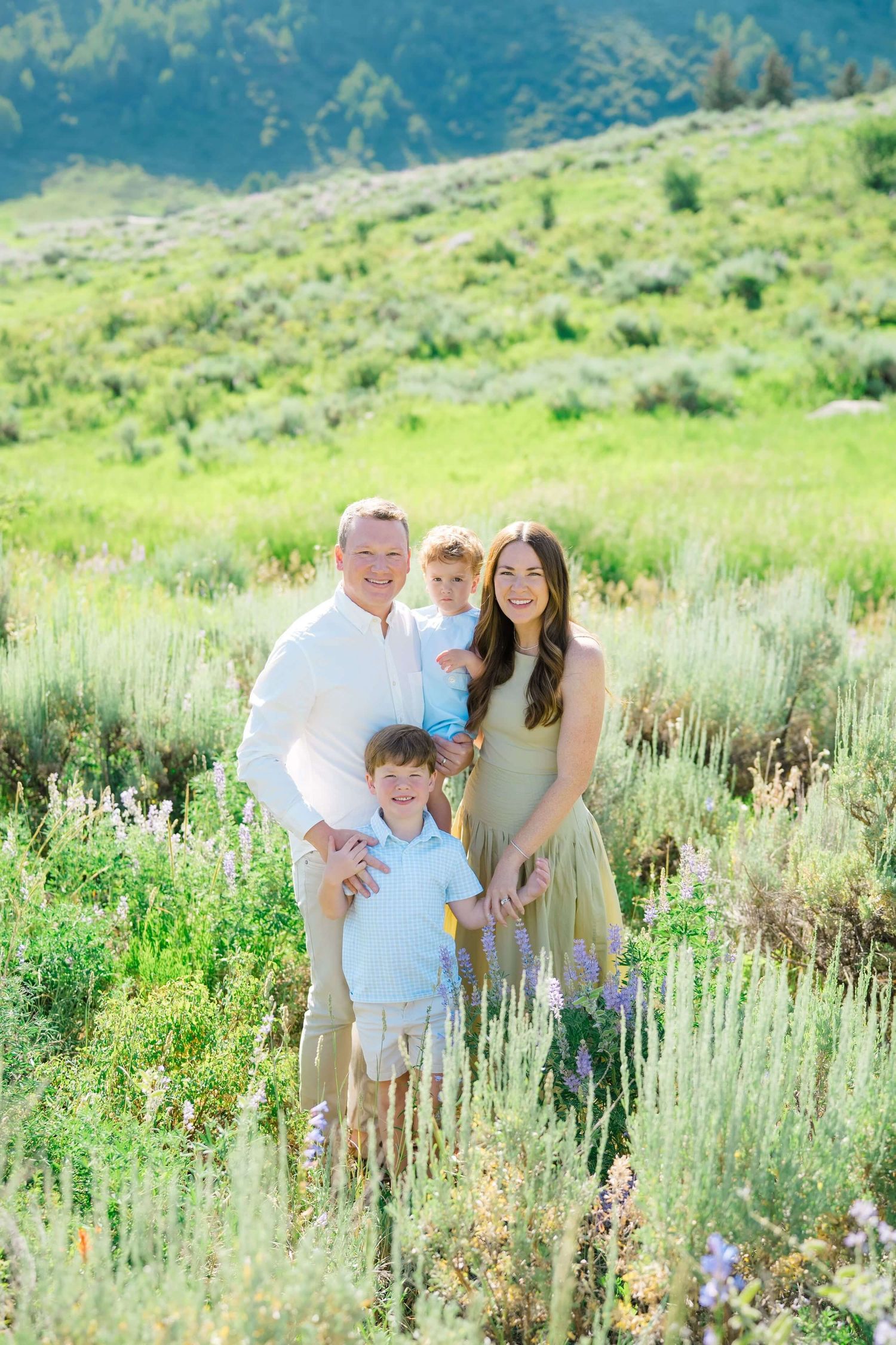 A family poses together in a scenic mountain meadow filled with tall grasses and wildflowers on a sunny day.