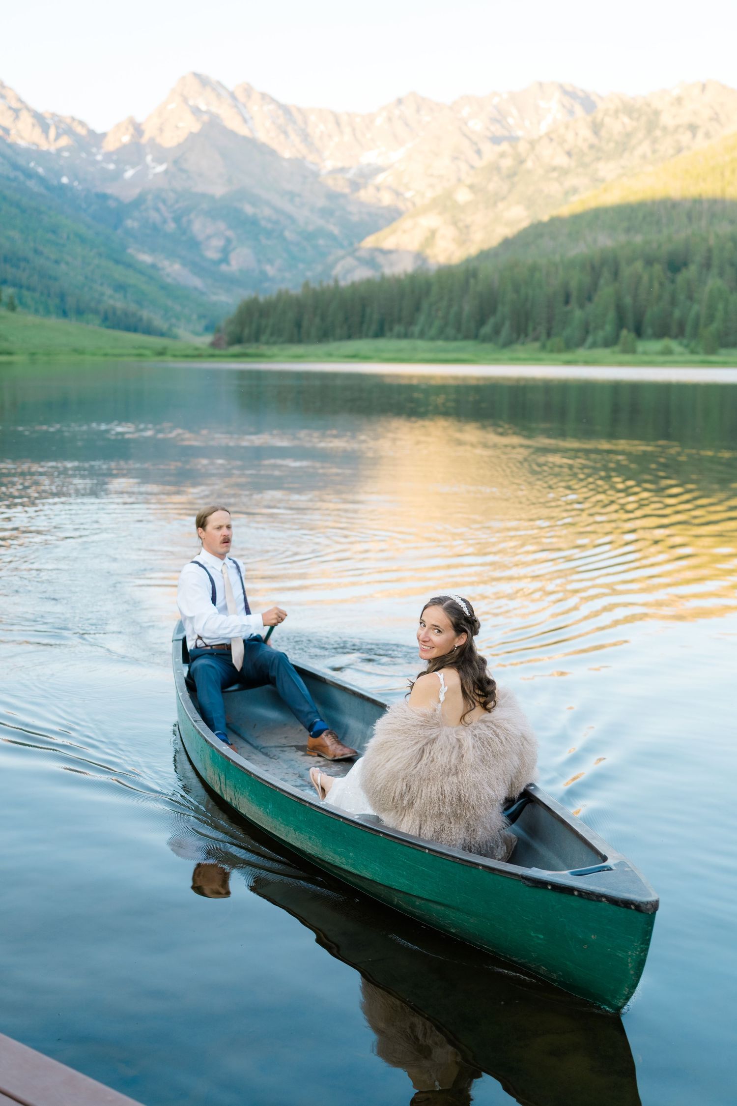Couple relaxes in green canoe on mountain lake during golden sunset with snow-capped peaks in background.