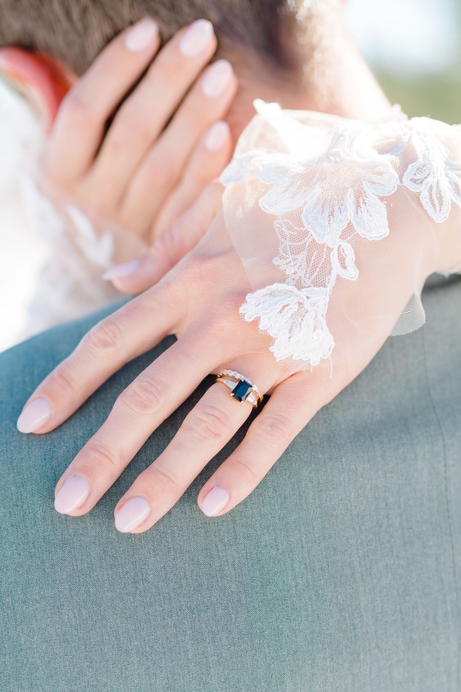 Close-up of delicate lace details and an engagement ring on a bride's hand against a gray background.