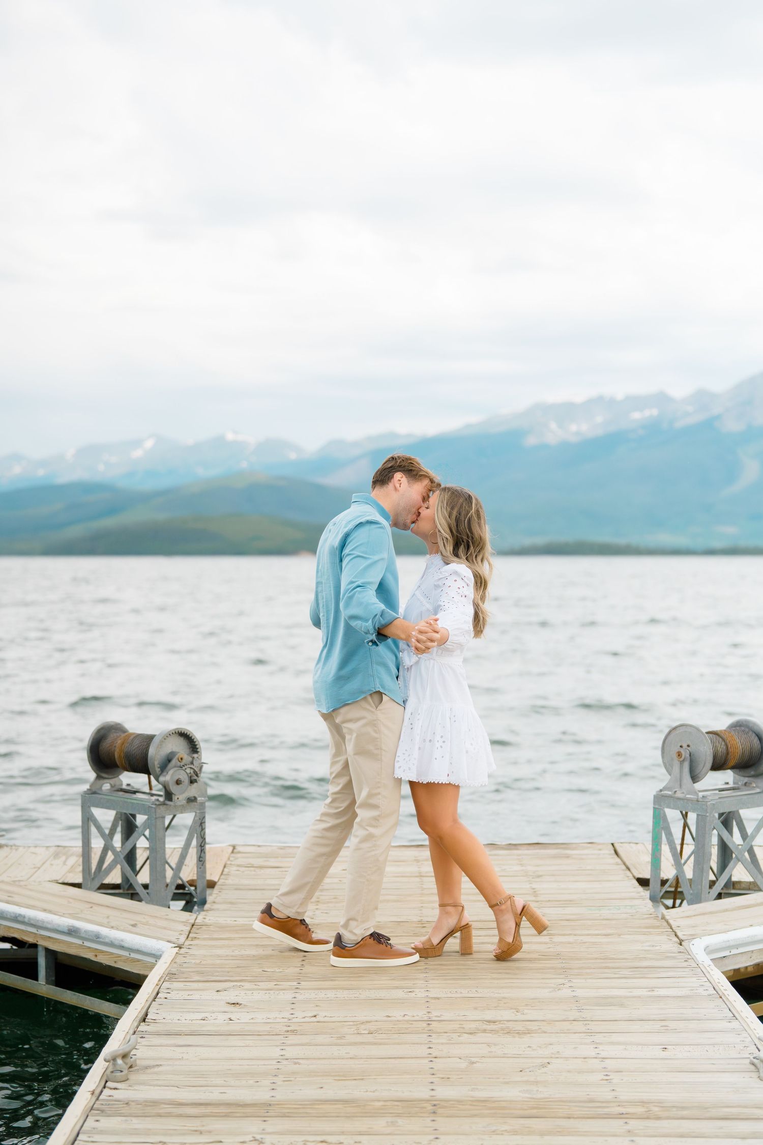 A couple embraces on a wooden dock overlooking a scenic mountain lake with cloudy skies in the background.