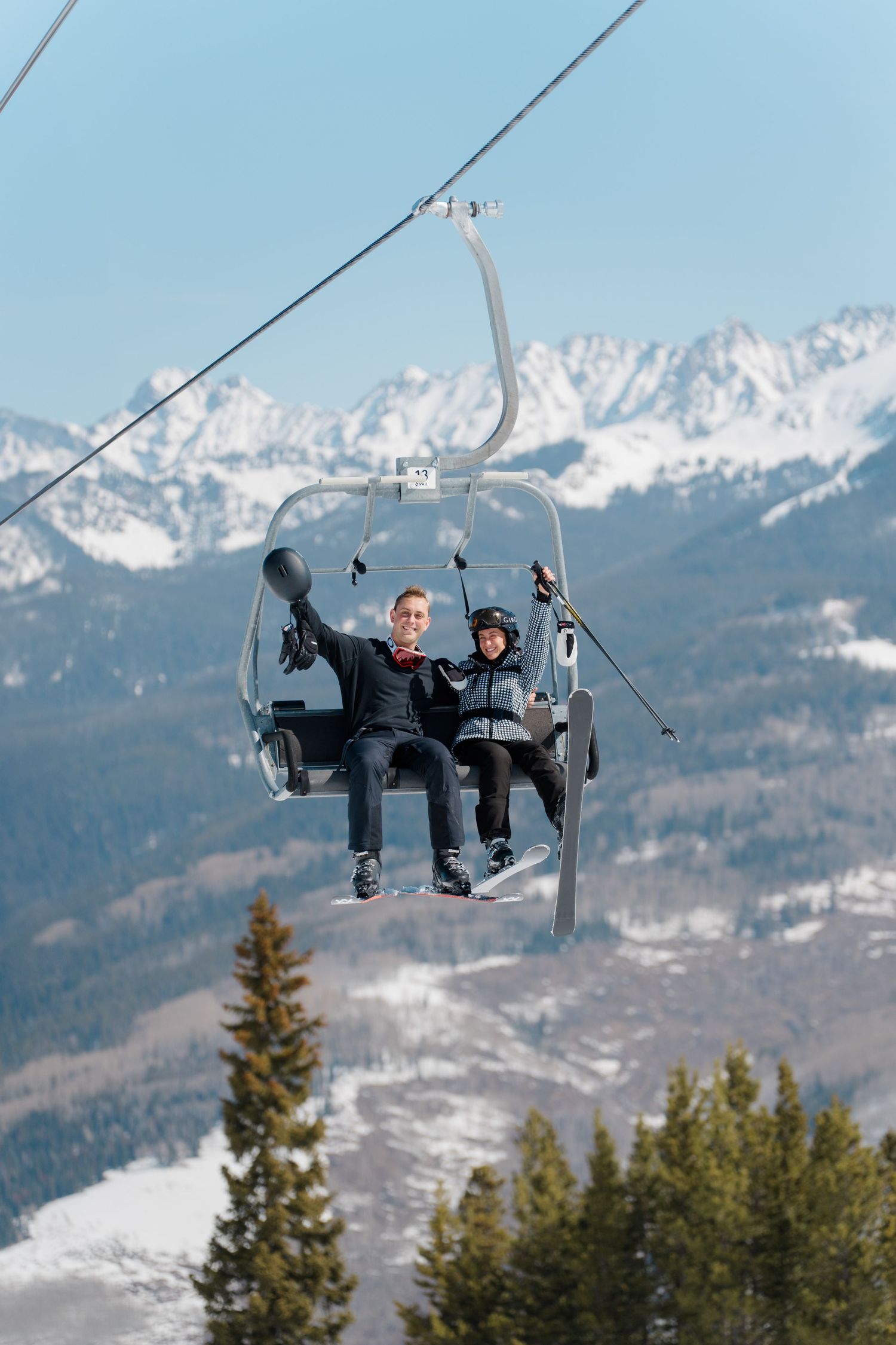 Two people playfully pose on a ski lift chair with snowy mountain peaks in the background.