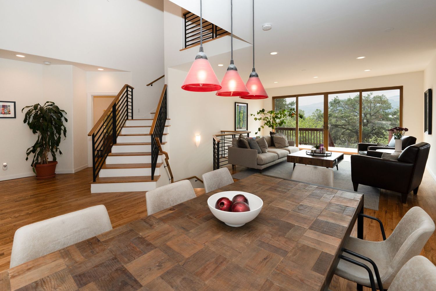 Modern open-plan living and dining area with red pendant lights, wood floors, staircase, and large windows overlooking greenery.