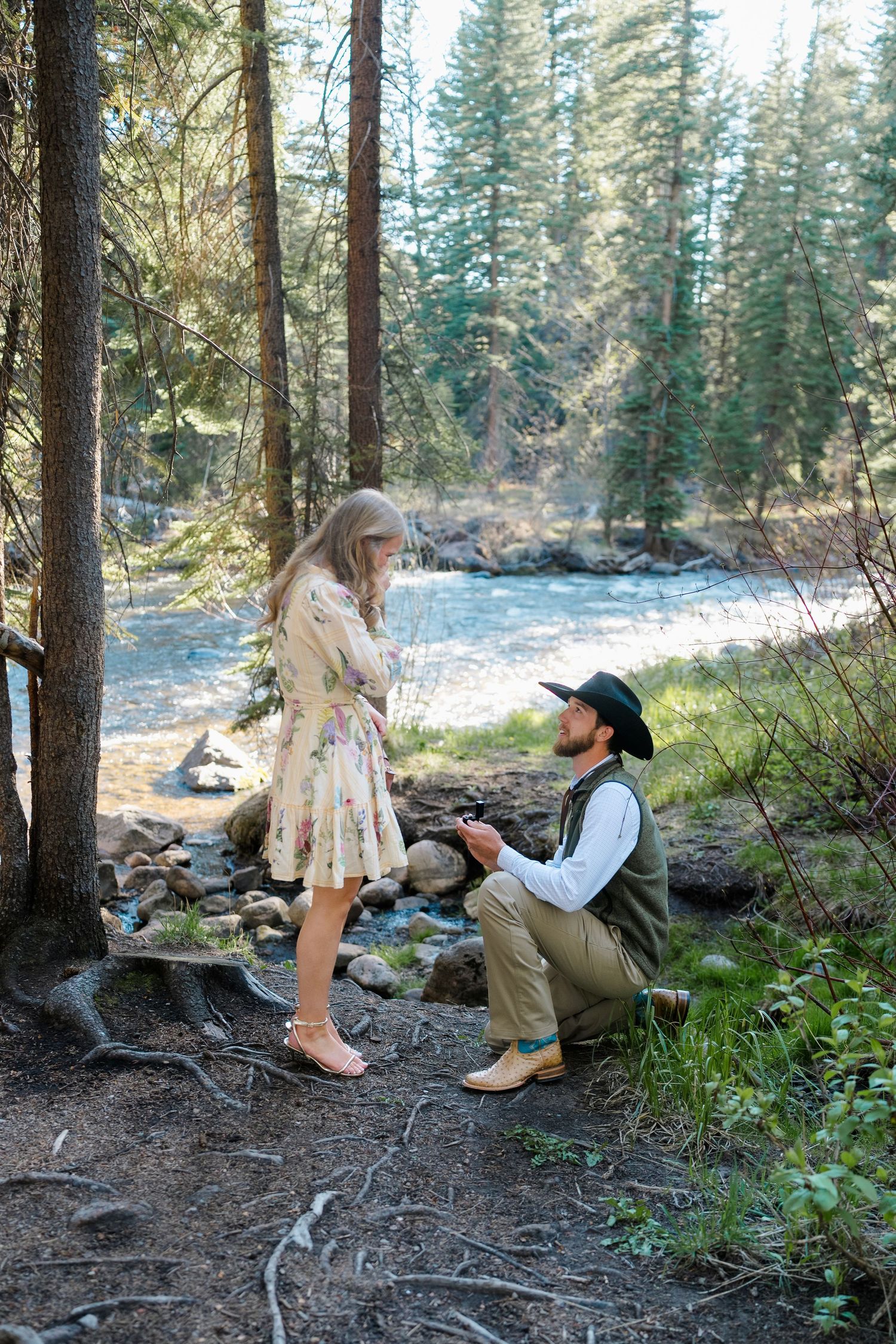 Man in cowboy hat proposes on one knee to woman in floral dress beside a forest river on a sunny day.