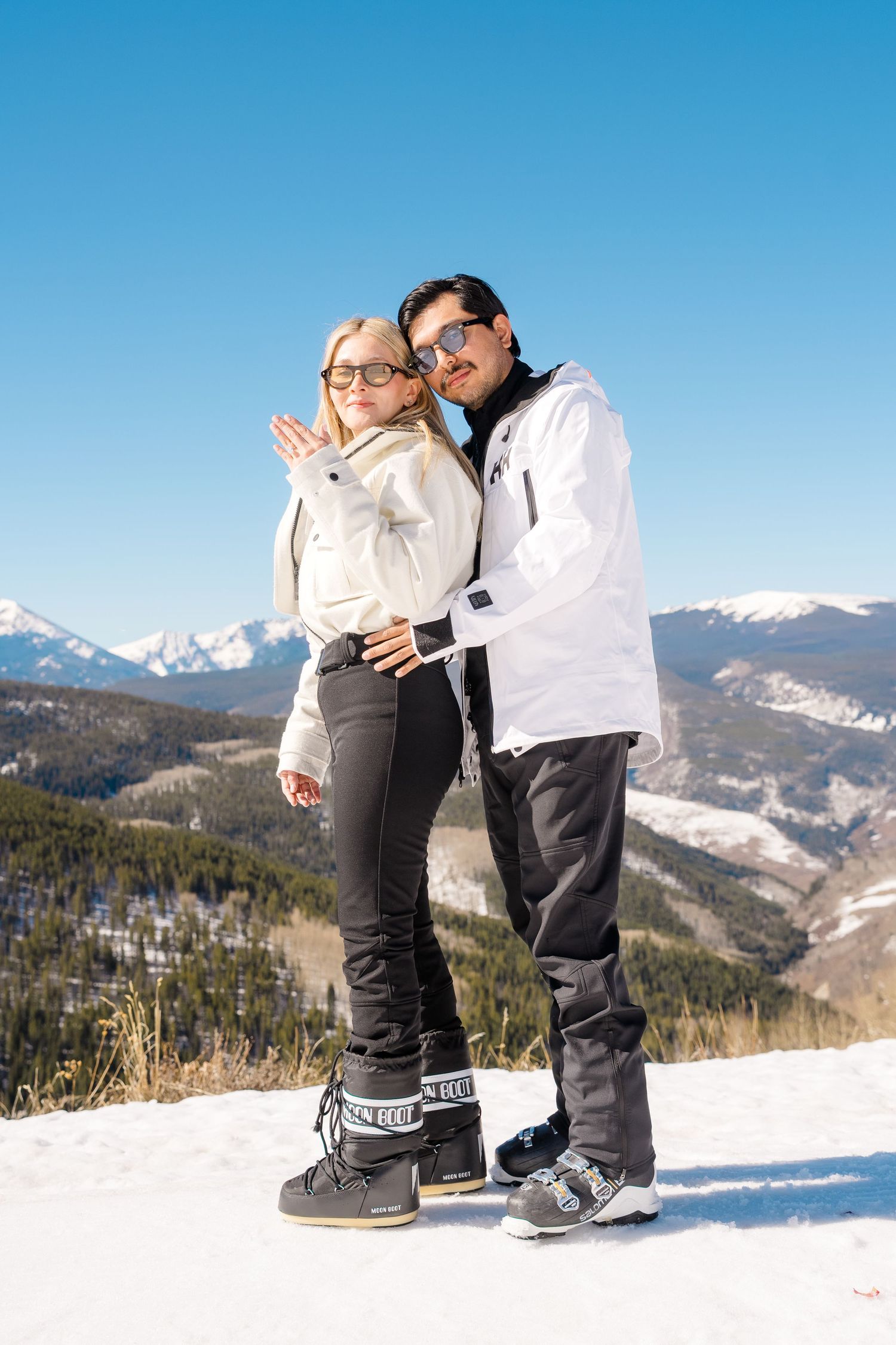 Happy couple posing together on snowy mountain, woman showing engagement ring with stunning alpine scenery.