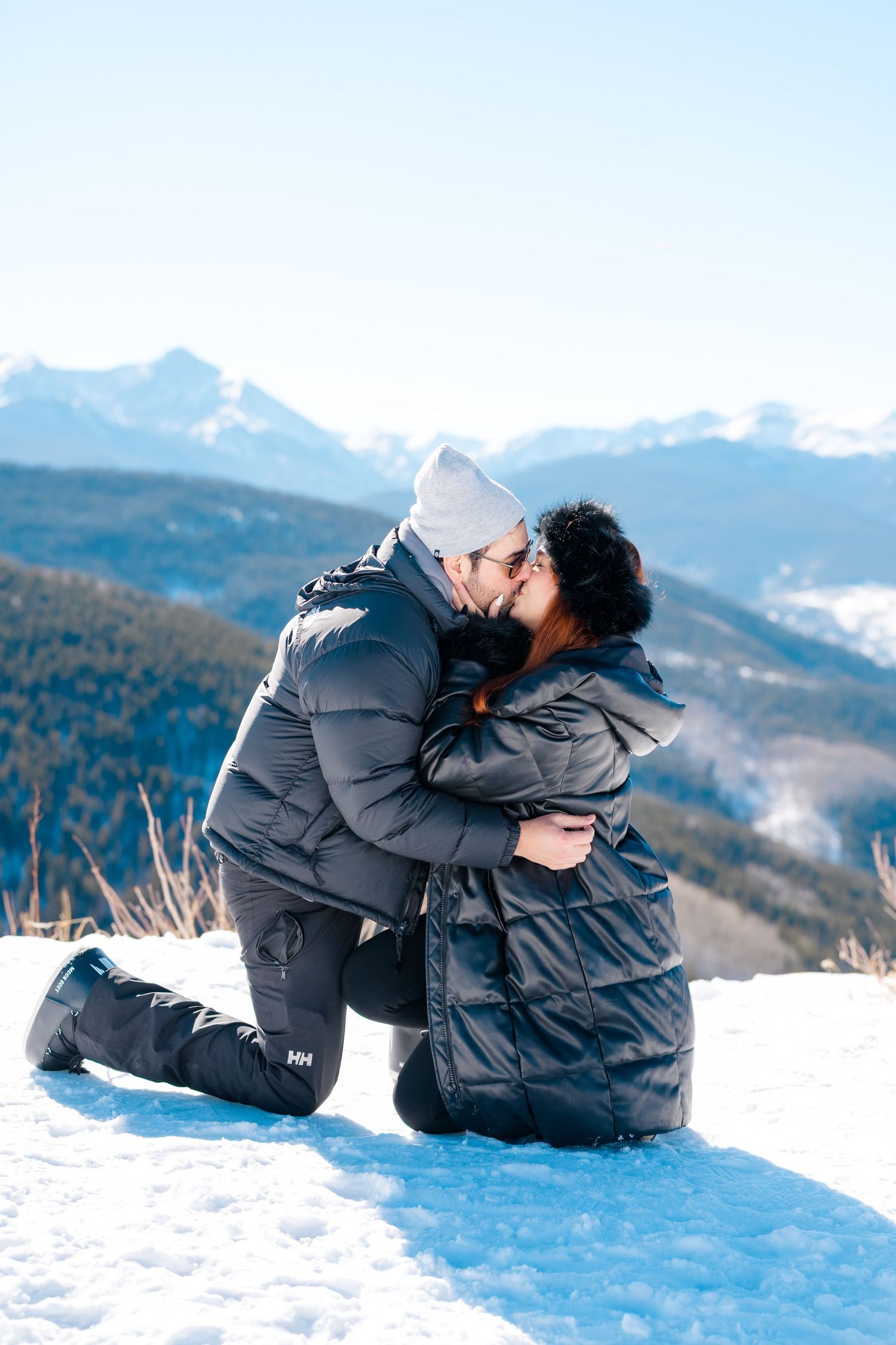 Two people in winter coats embrace while sitting in snow with mountain peaks visible in the background at vail proposal.