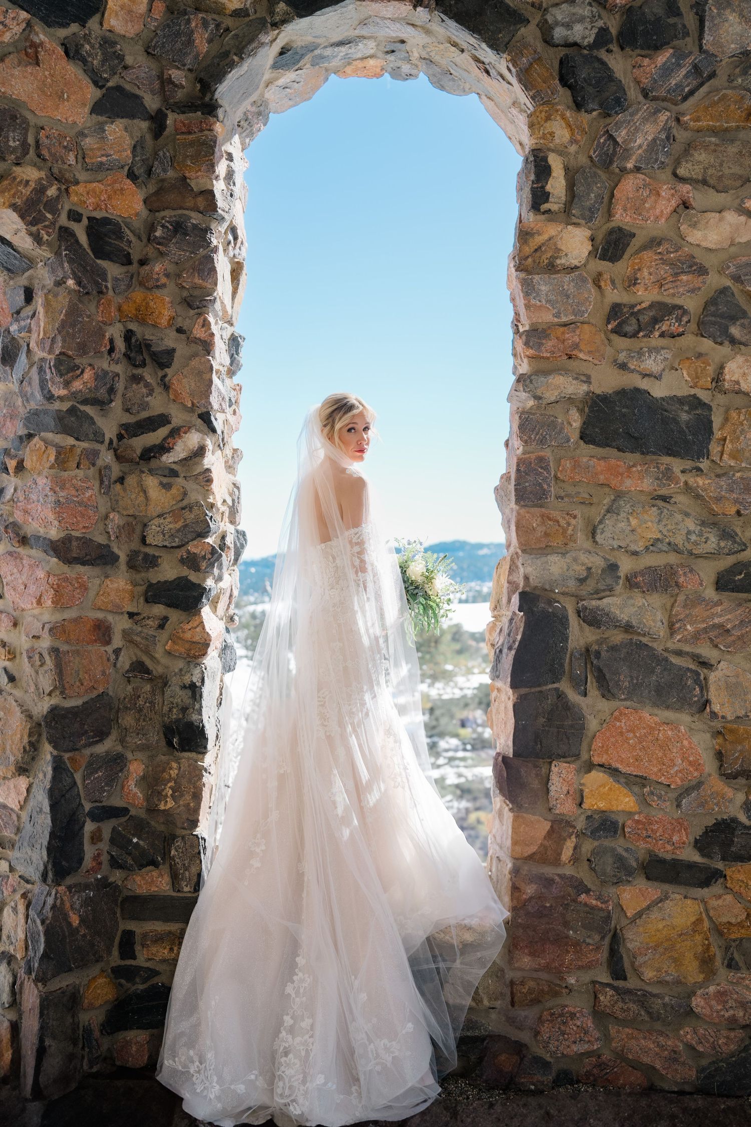 Bride in lace wedding gown and veil stands in stone arch window holding bouquet with blue sky backdrop.